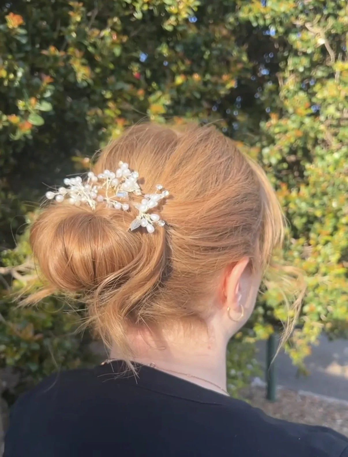 A woman with red hair styled in a bun with a pearl and silver decorative hairpiece, standing outdoors with green foliage in the background.
