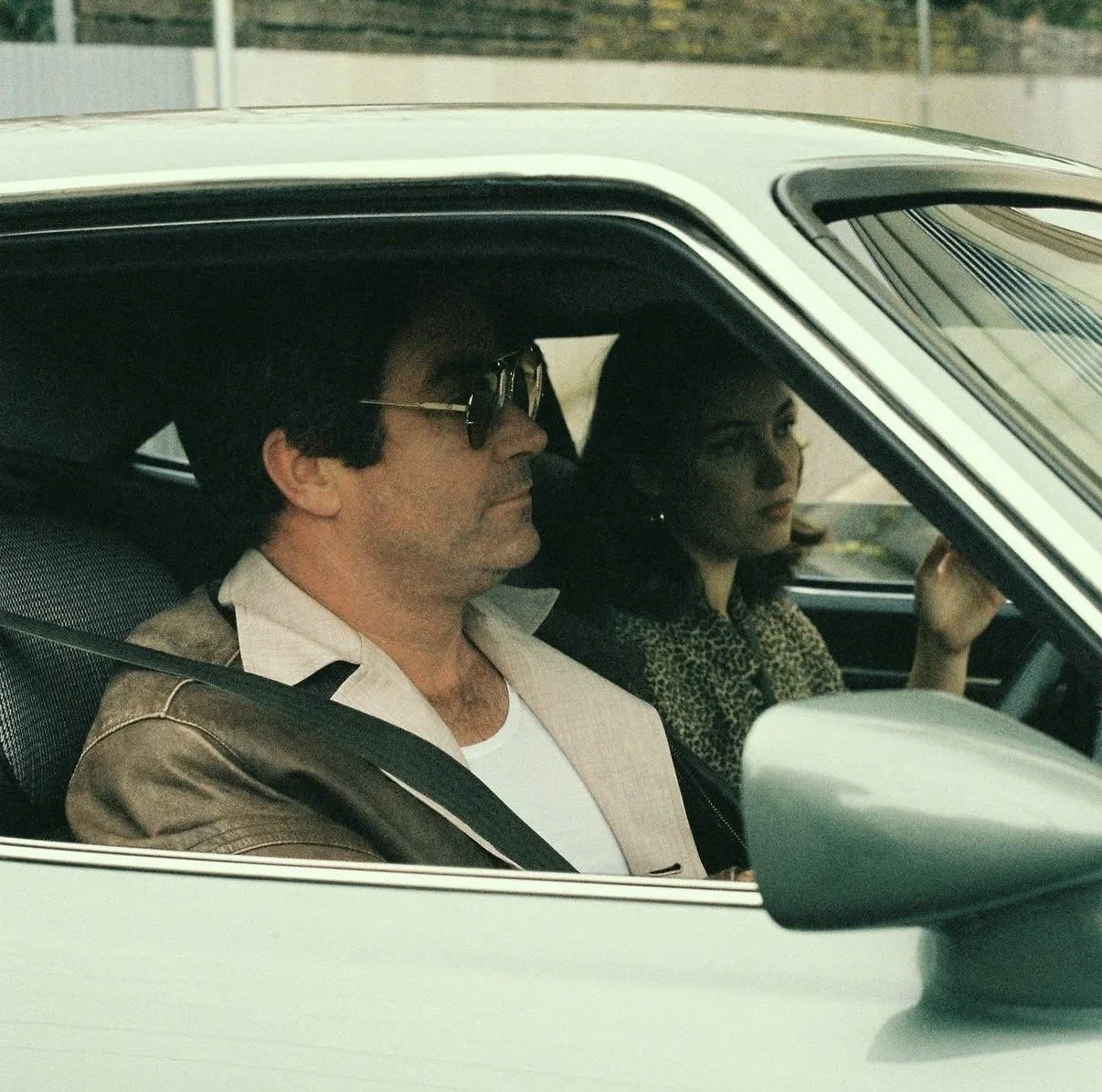 A man with dark hair, wearing sunglasses, a white shirt, and a beige jacket, is sitting in the driver's seat of a vintage white car. A woman with dark hair, wearing a leopard print top, is sitting in the passenger seat, looking out the window.