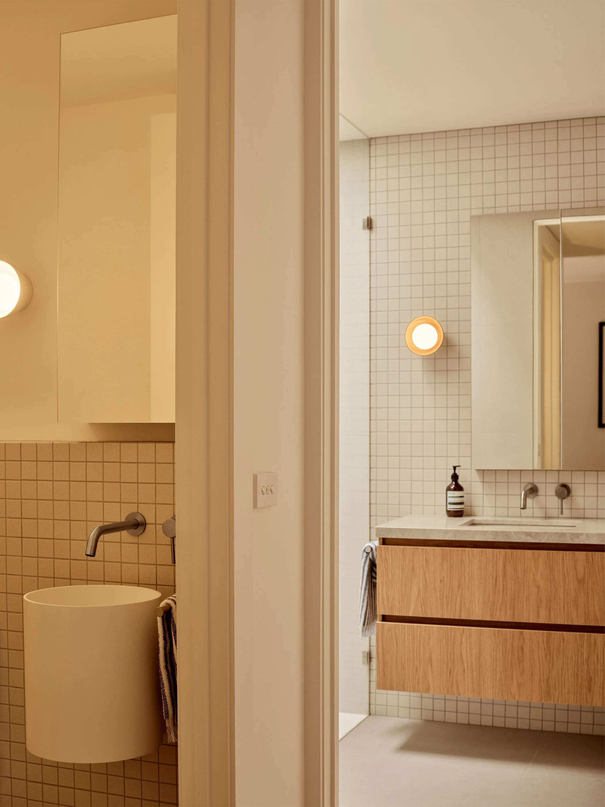 Bathroom with a beige tiled wall and modern wooden vanity with a white countertop. A soap dispenser is on the sink, and a mirror is mounted above. The bathroom has a round ceiling light and a cream-colored cylindrical trash can with a striped towel h