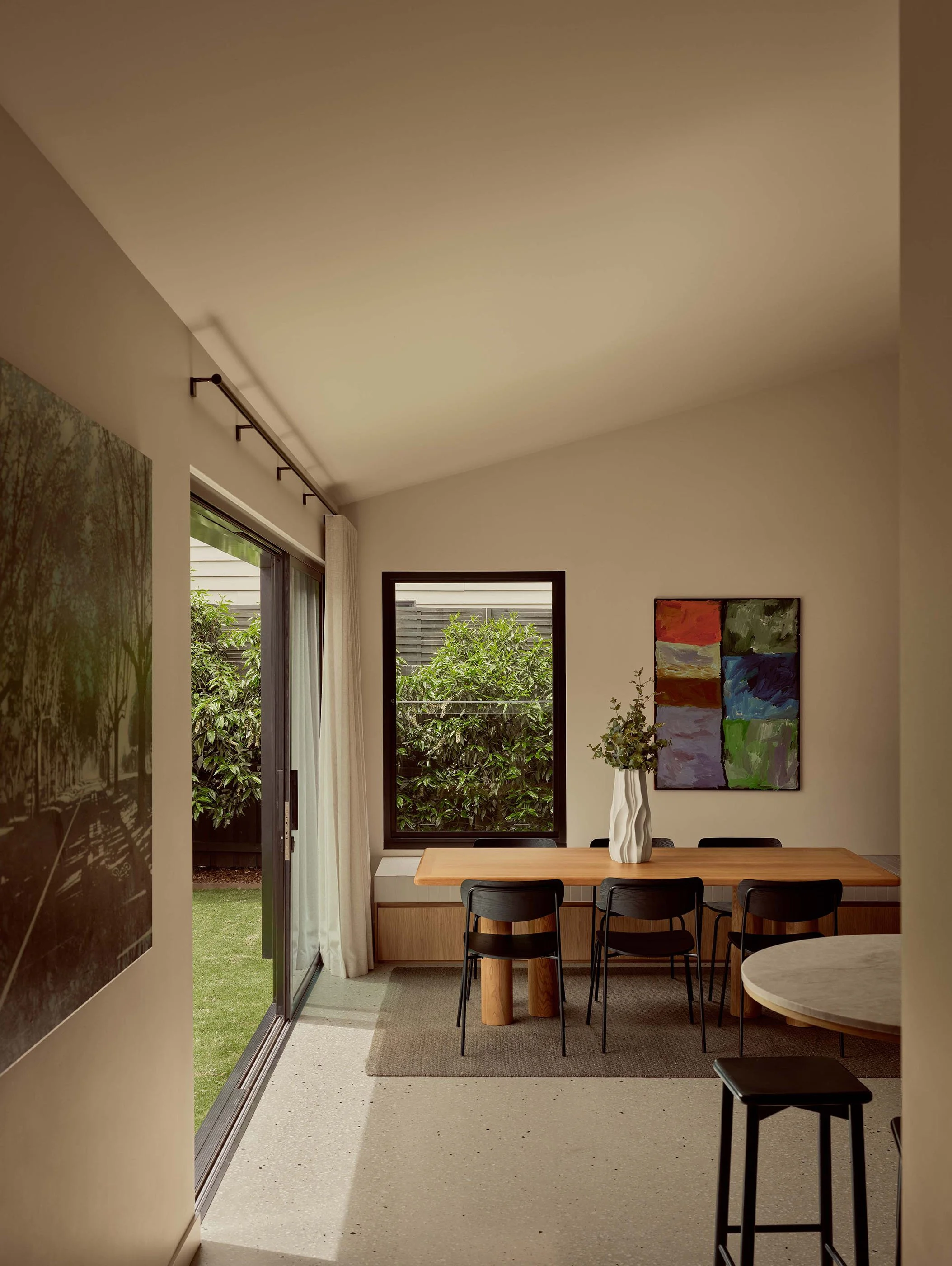 Interior of a dining room with a wooden table, black chairs, a large abstract painting, a white vase with greenery, and a sliding glass door leading to a backyard with bushes.
