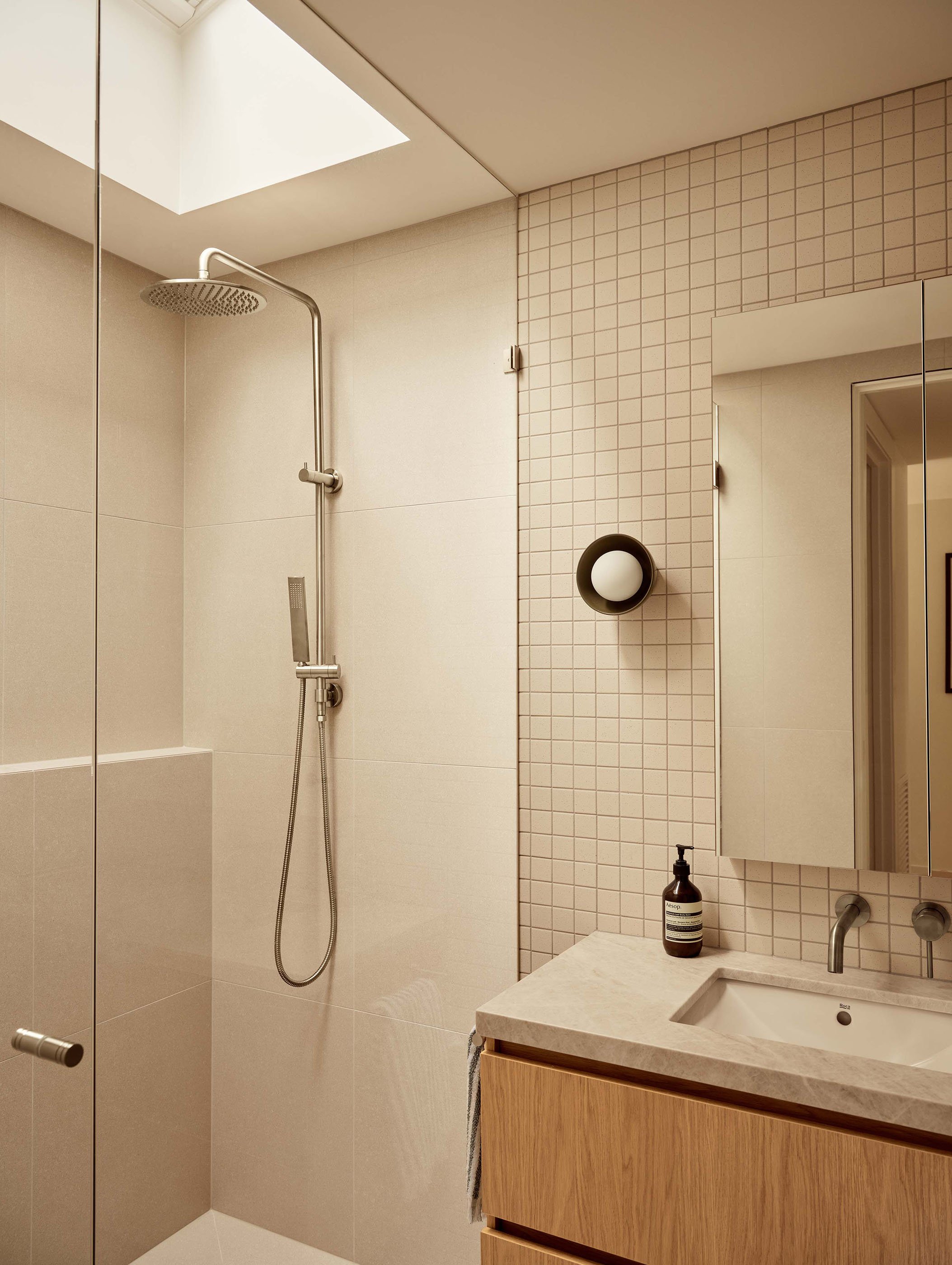 Modern bathroom with a walk-in shower, beige tiles, a skylight, a mirror, a wooden vanity with a white sink, a black pump soap dispenser, and a round wall light.