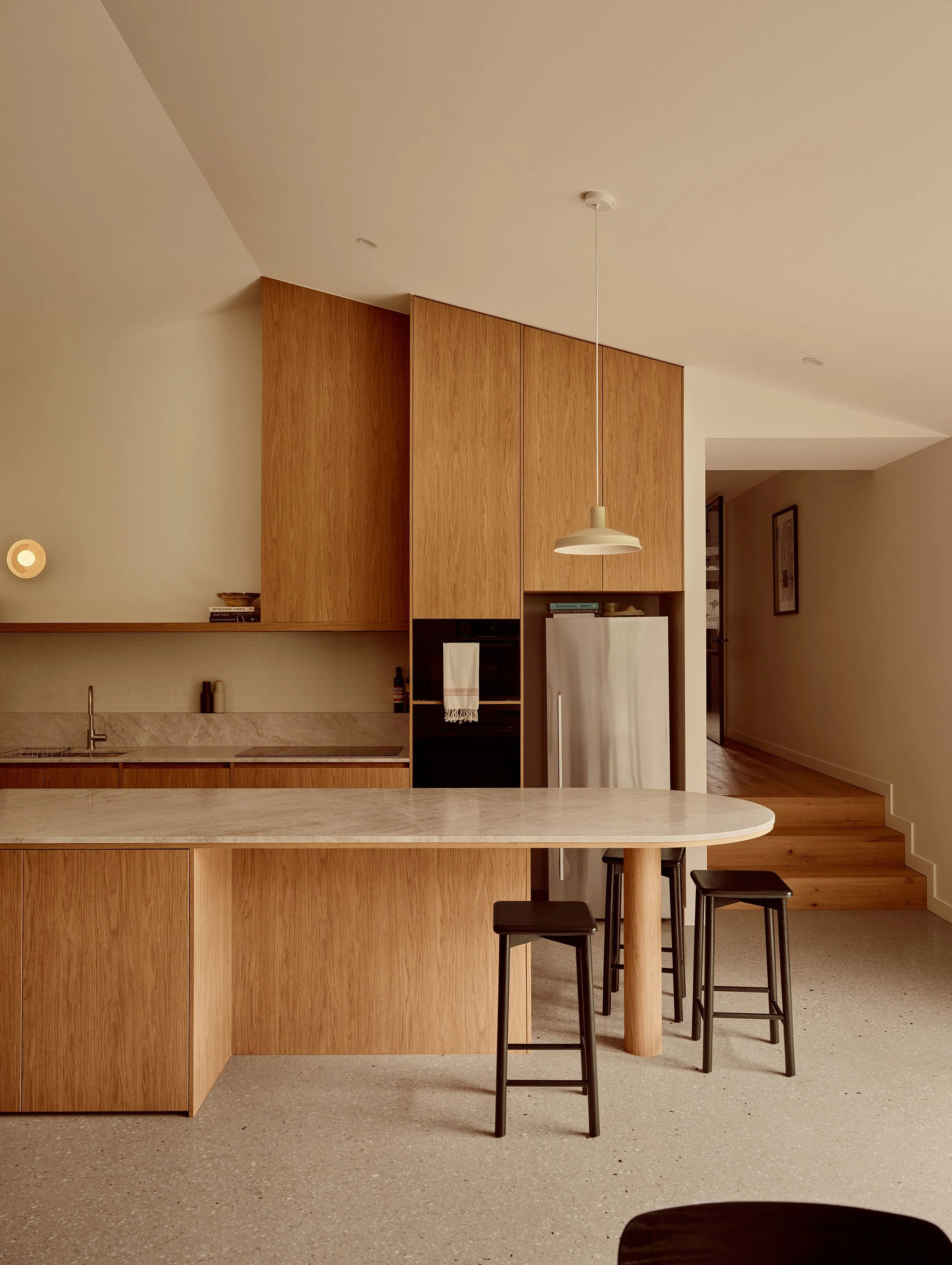 Modern kitchen with wood cabinets, marble island, black stools, white refrigerator, pendant light, and continuation of living space hallway.