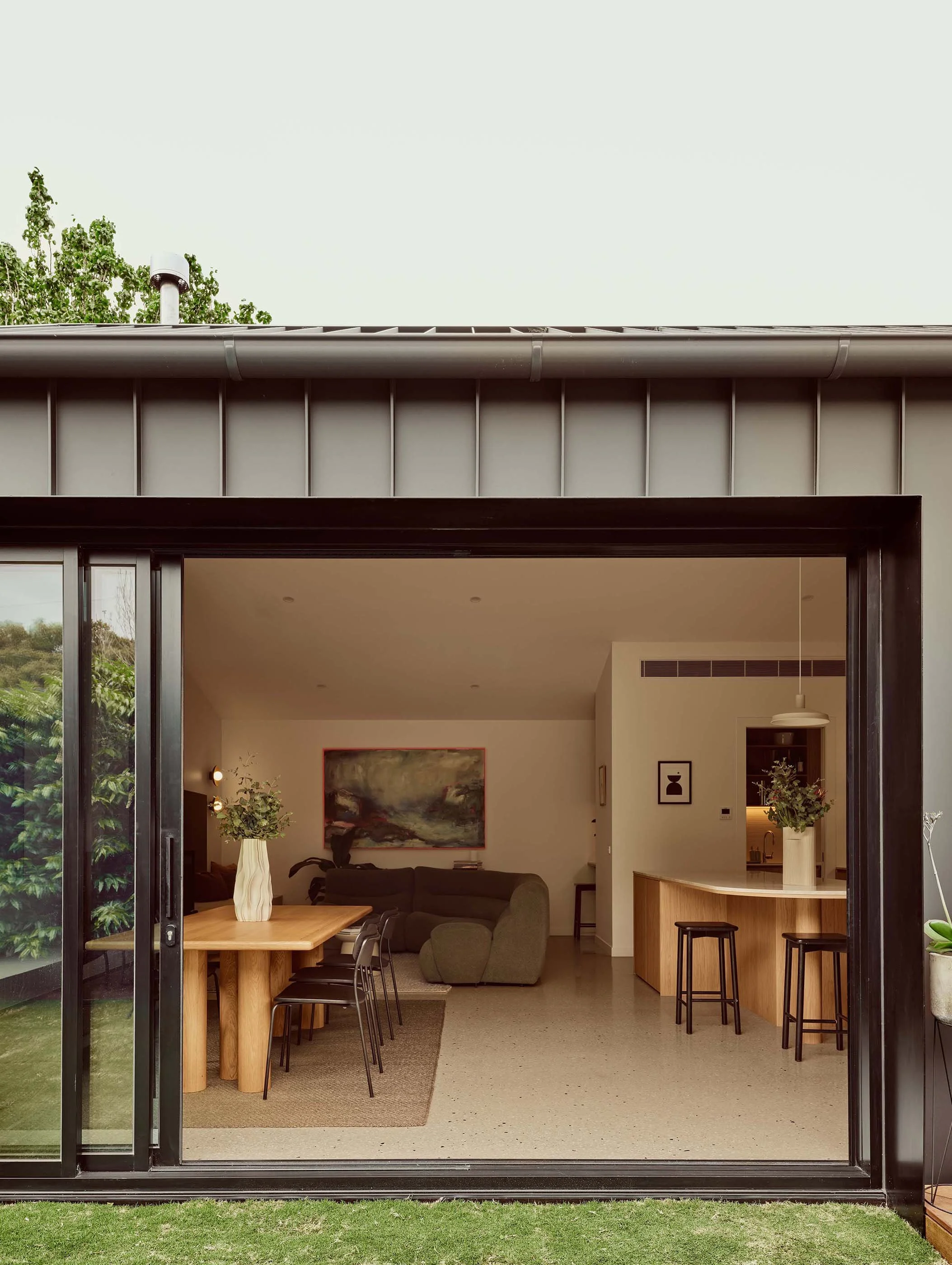 A modern living room with large glass sliding doors, a dining table with black chairs, a gray sectional sofa, artwork on the wall, and a kitchen area with bar stools and a light fixture.