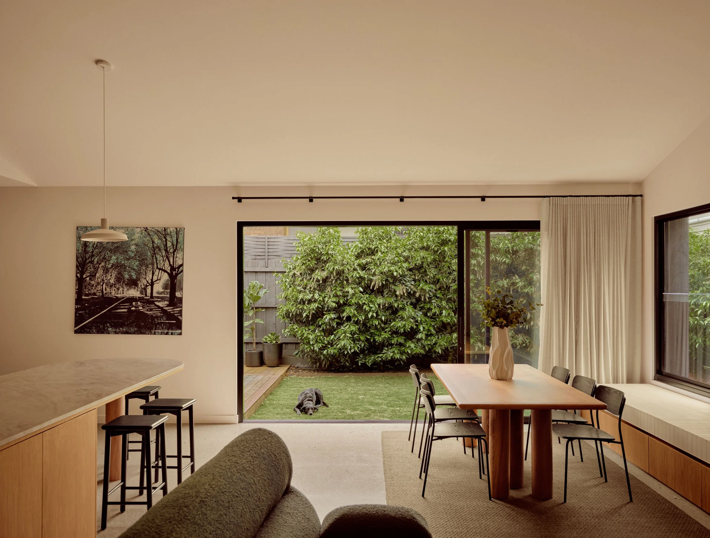 A living room with large glass sliding doors opening to a backyard with a dog lying on the grass. The room has a wooden table with black chairs, a hanging pendant light, and a black and white wall art depicting a tree-lined street.