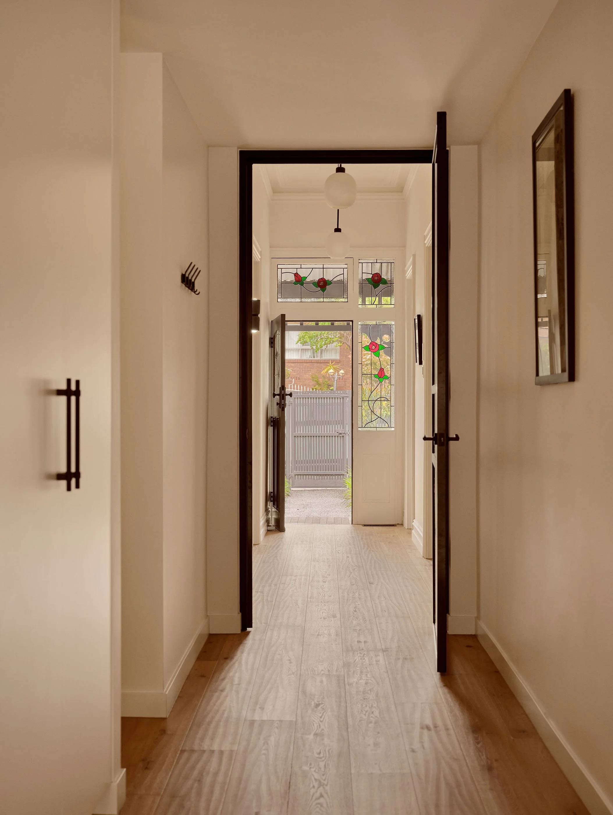 A hallway with wooden flooring, white walls, and a door at the end with decorative stained glass panels featuring red roses and green leaves. There are two ceiling lights hanging and a mirror on the right wall.