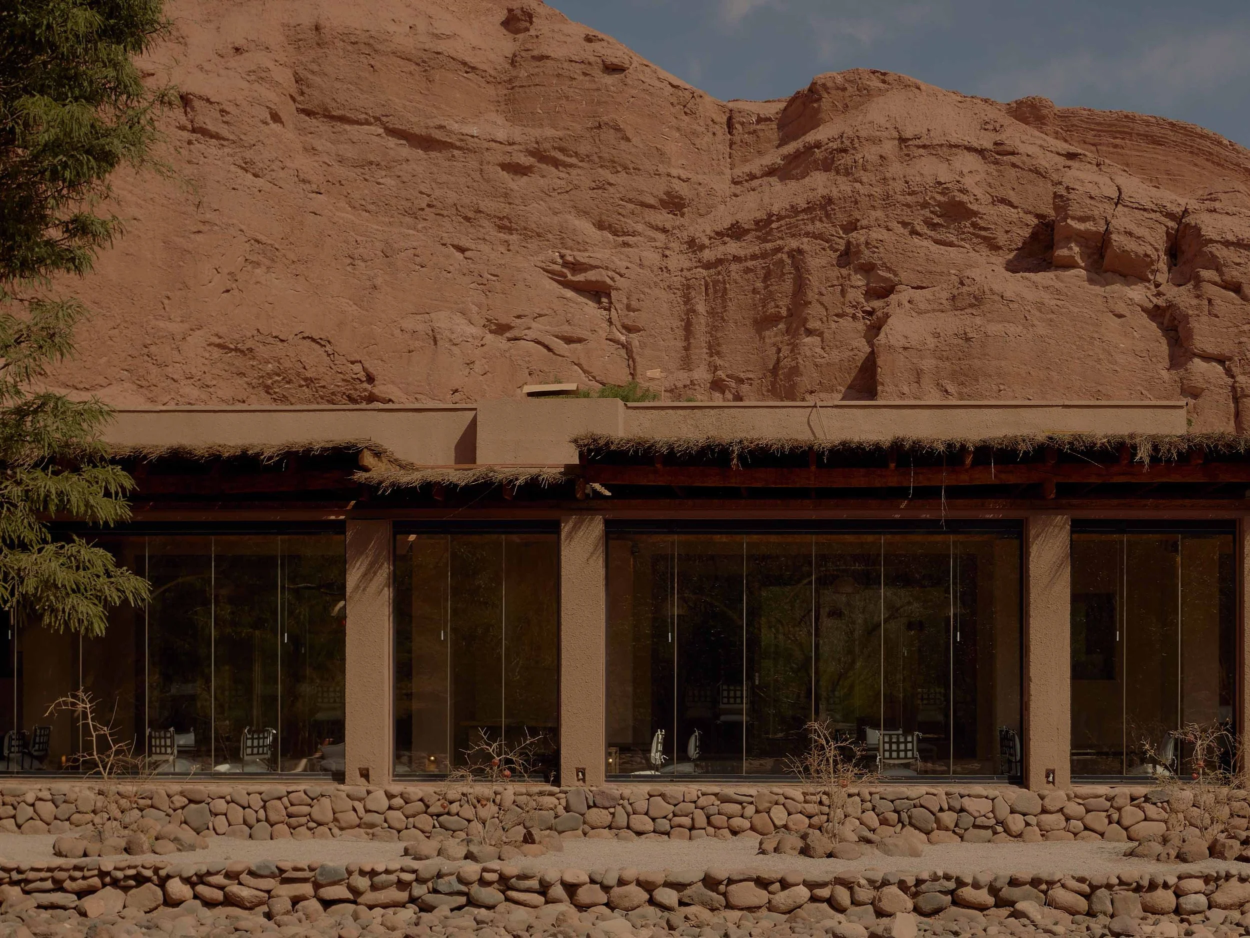 A modern building with large glass windows and a thatched roof, set against a rocky desert landscape with reddish-brown cliffs and sparse vegetation.