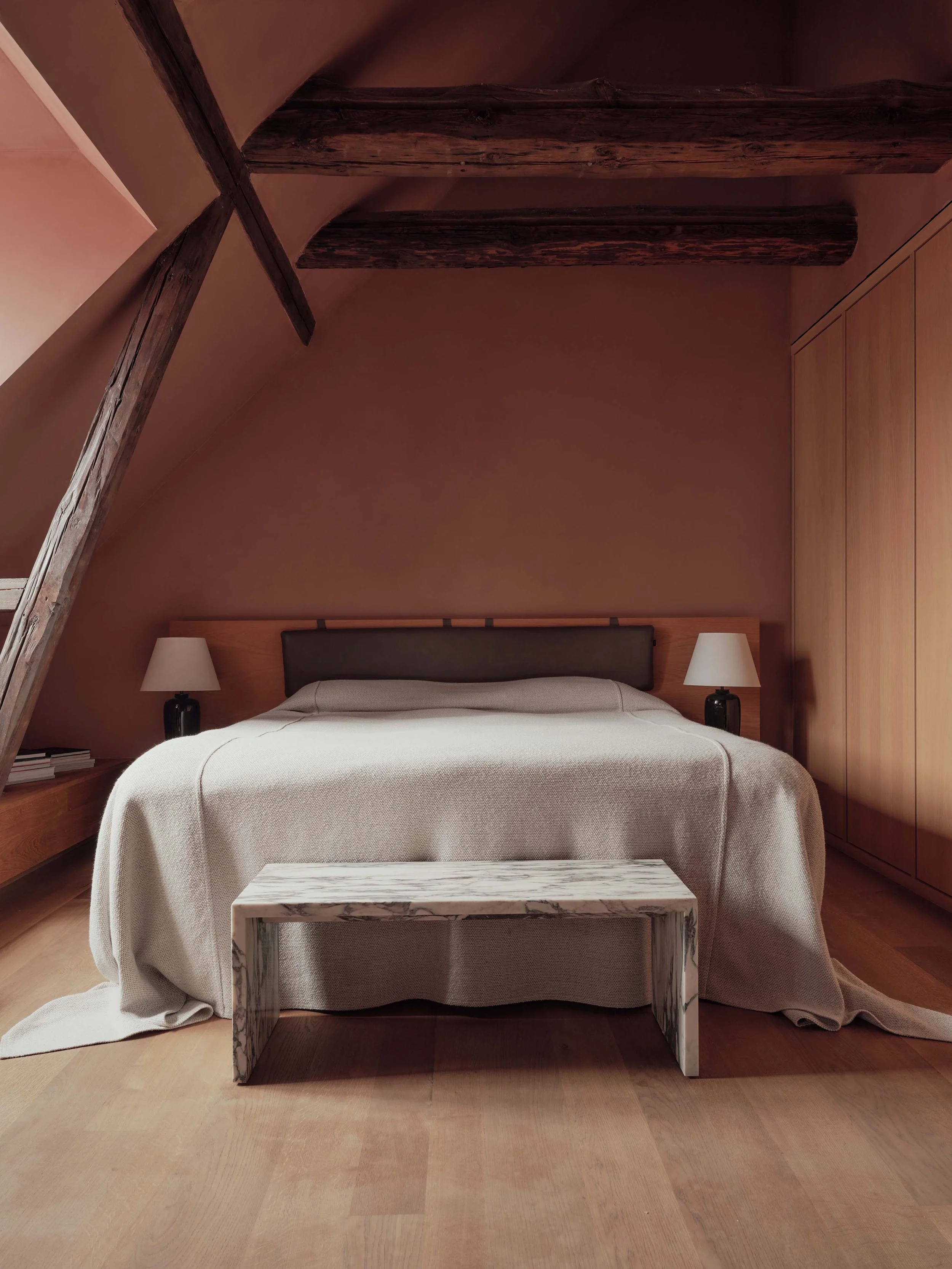 A bedroom with a bed covered in a beige blanket, two black bedside lamps, and a small marble table in front of the bed. The wall behind the bed is wood-paneled and there are exposed wooden beams on the ceiling.