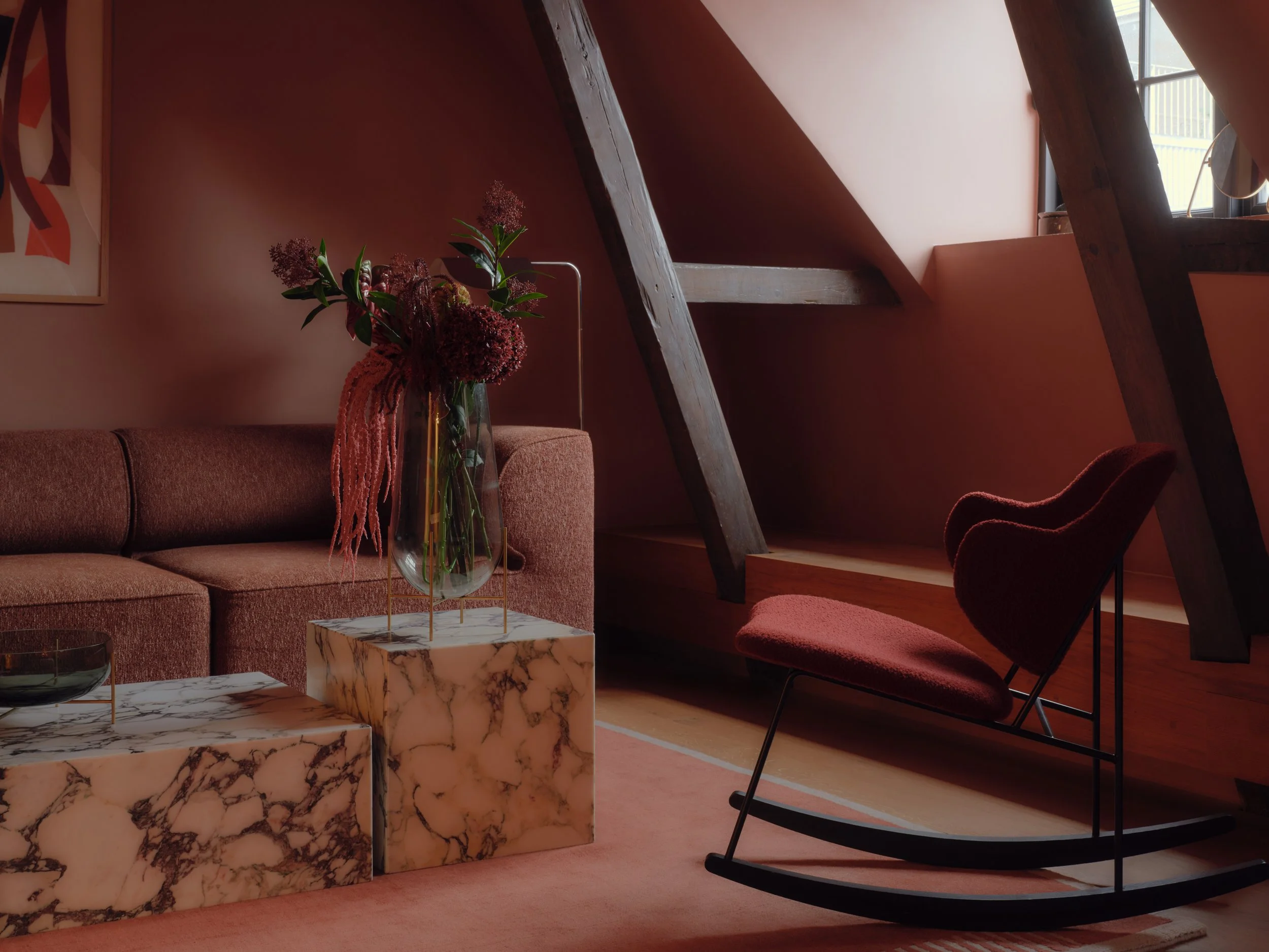 A cozy living room with a pinkish wall, a maroon sofa with patterned cushions, a marble-topped coffee table, a glass vase with pink flowers, and a black rocking chair near a window with wooden beams overhead.