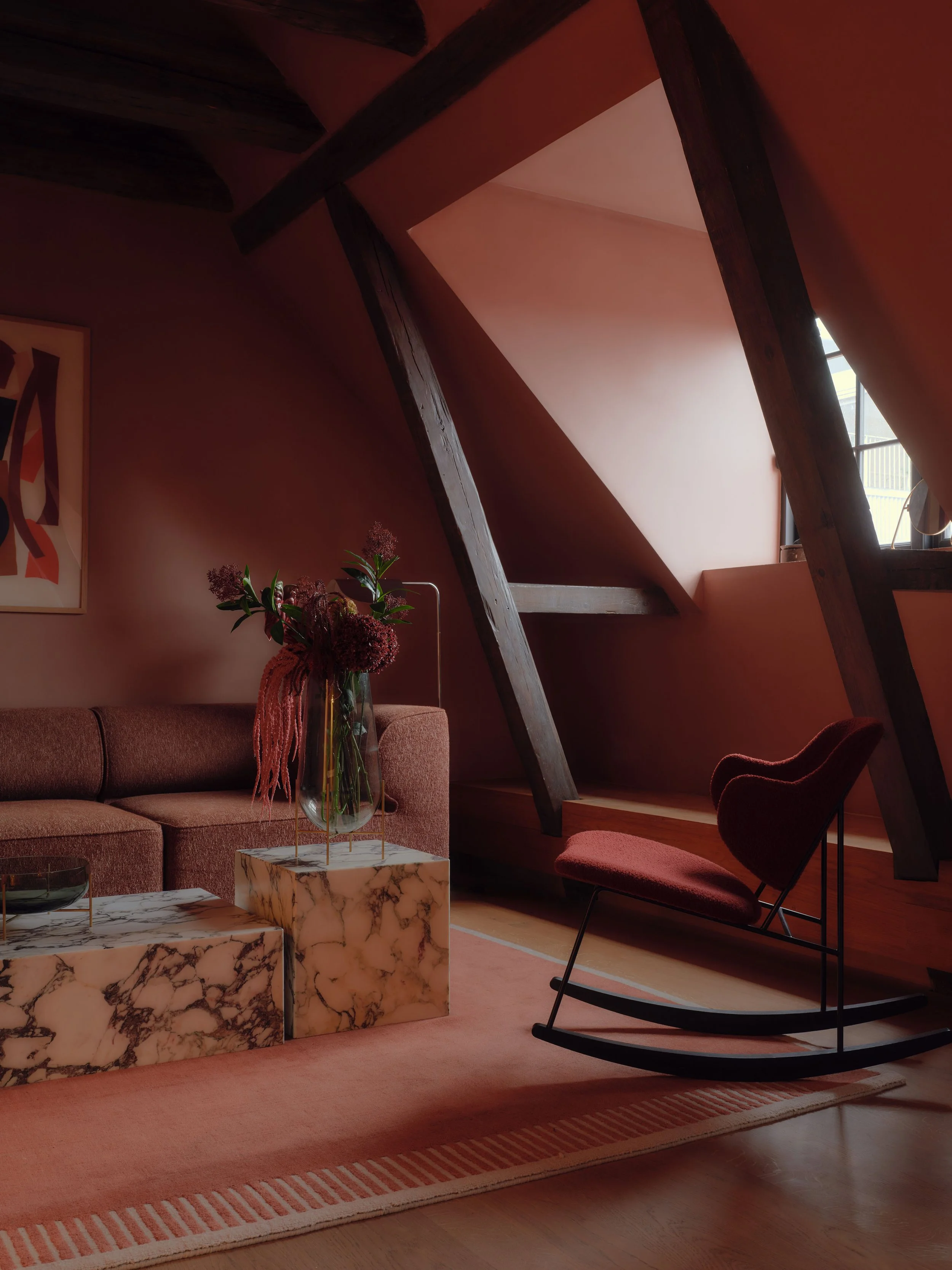 A cozy living room with sloped ceiling and large window, featuring a pink sofa, dark wood beams, a marble coffee table, a rocking chair, and a vase with pink flowers.