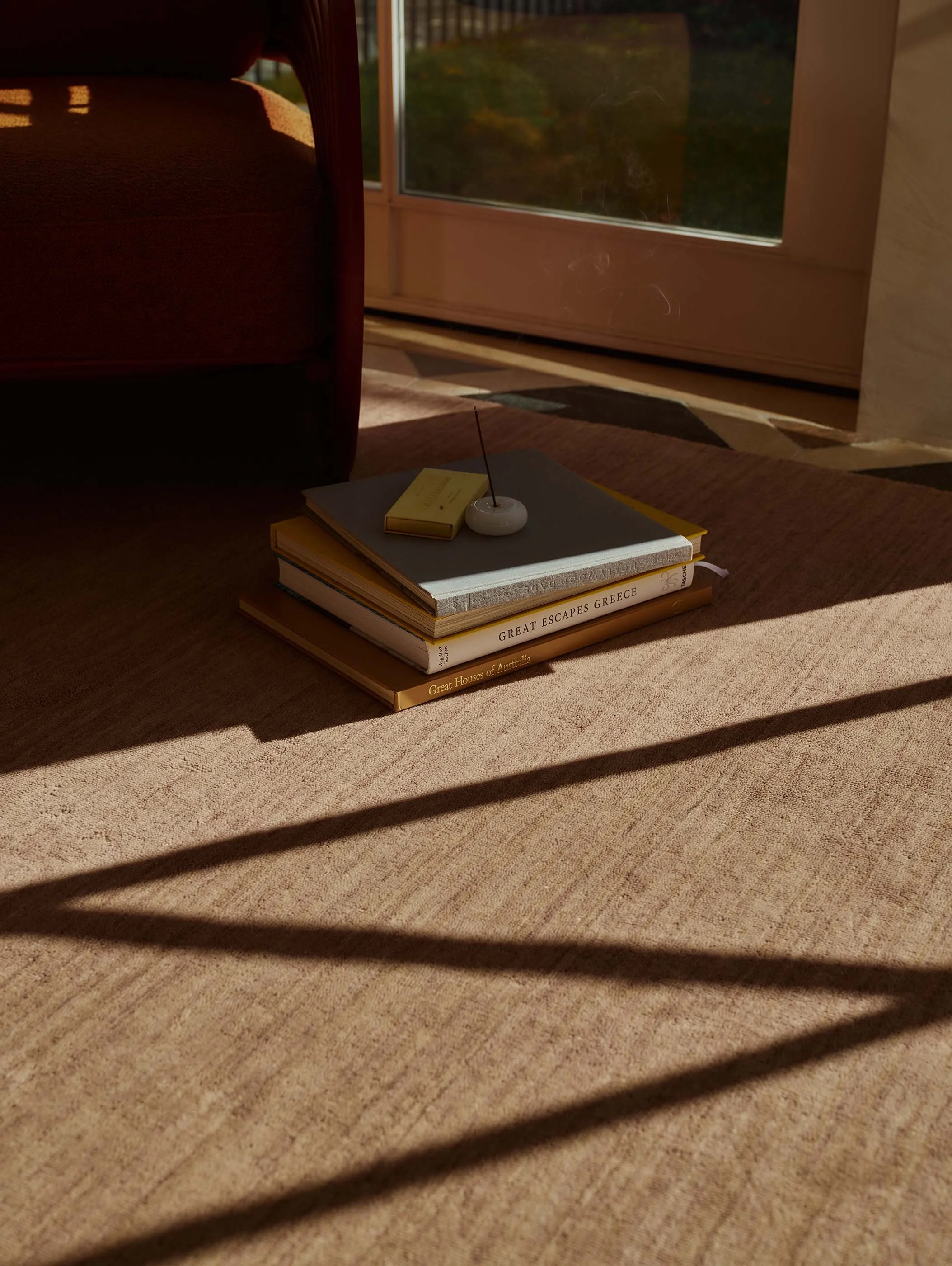 A close-up of a stack of books on a carpeted floor near a window, with shadows cast on the carpet from window blinds, and sunlight illuminating the books and part of the floor.