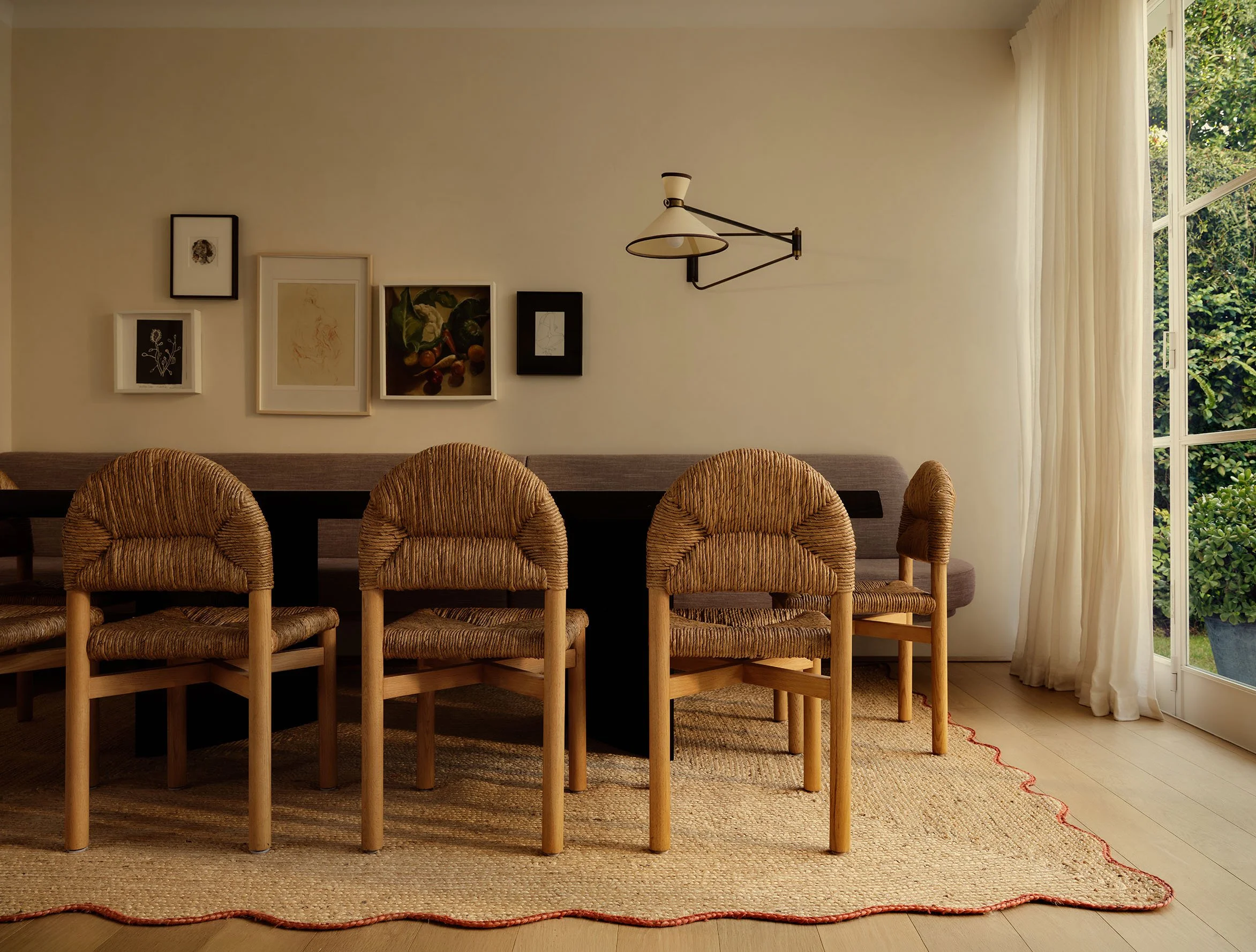 A cozy dining area with five rattan chairs around a dark wooden table, decorative framed artwork on a beige wall, a modern wall-mounted lamp, large windows with sheer curtains, and a textured beige rug on a light wooden floor.
