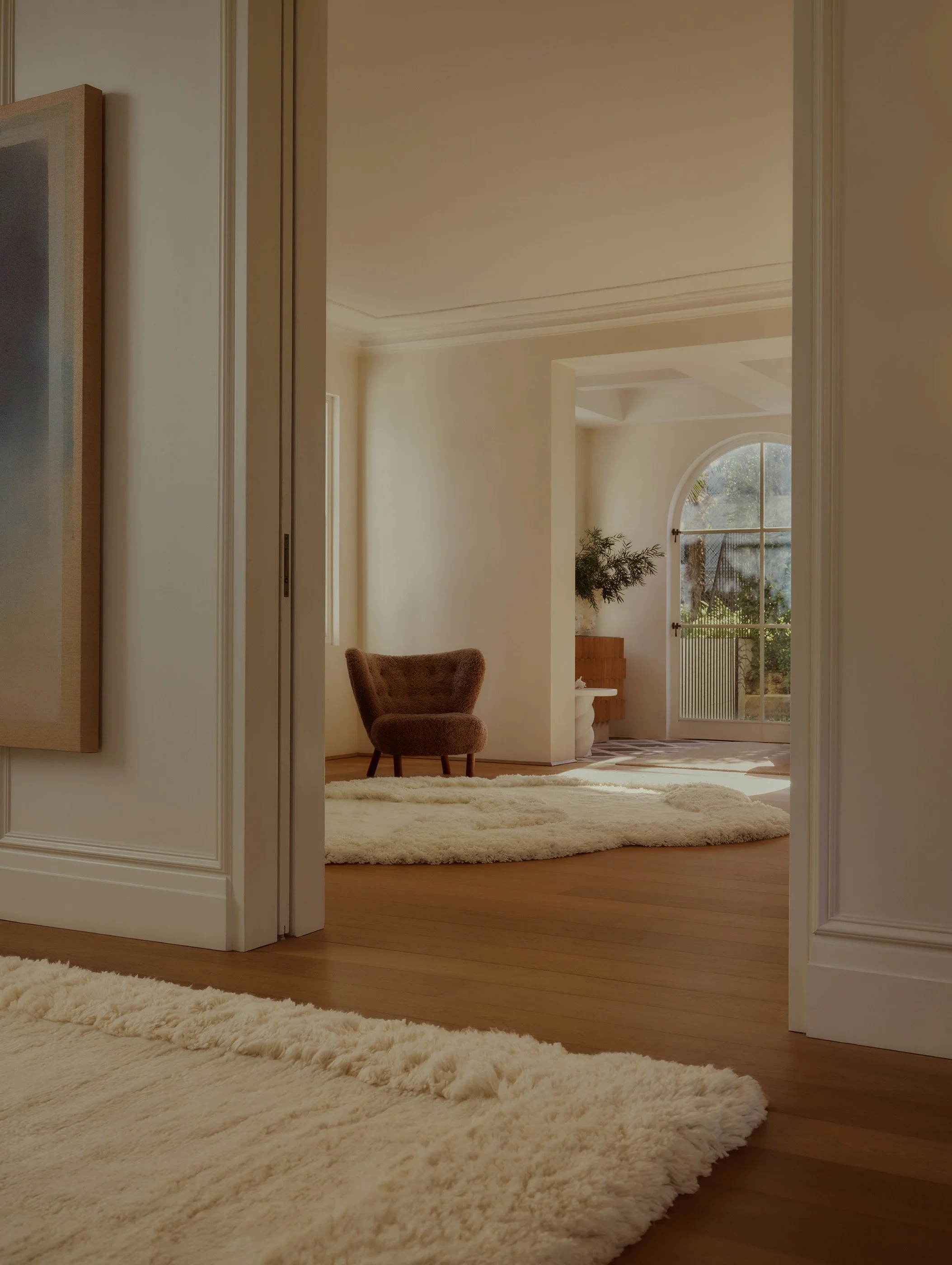 View of a cozy living room with a large, cream-colored shag rug, a plush armchair, a window with natural light, and a decorative fireplace.