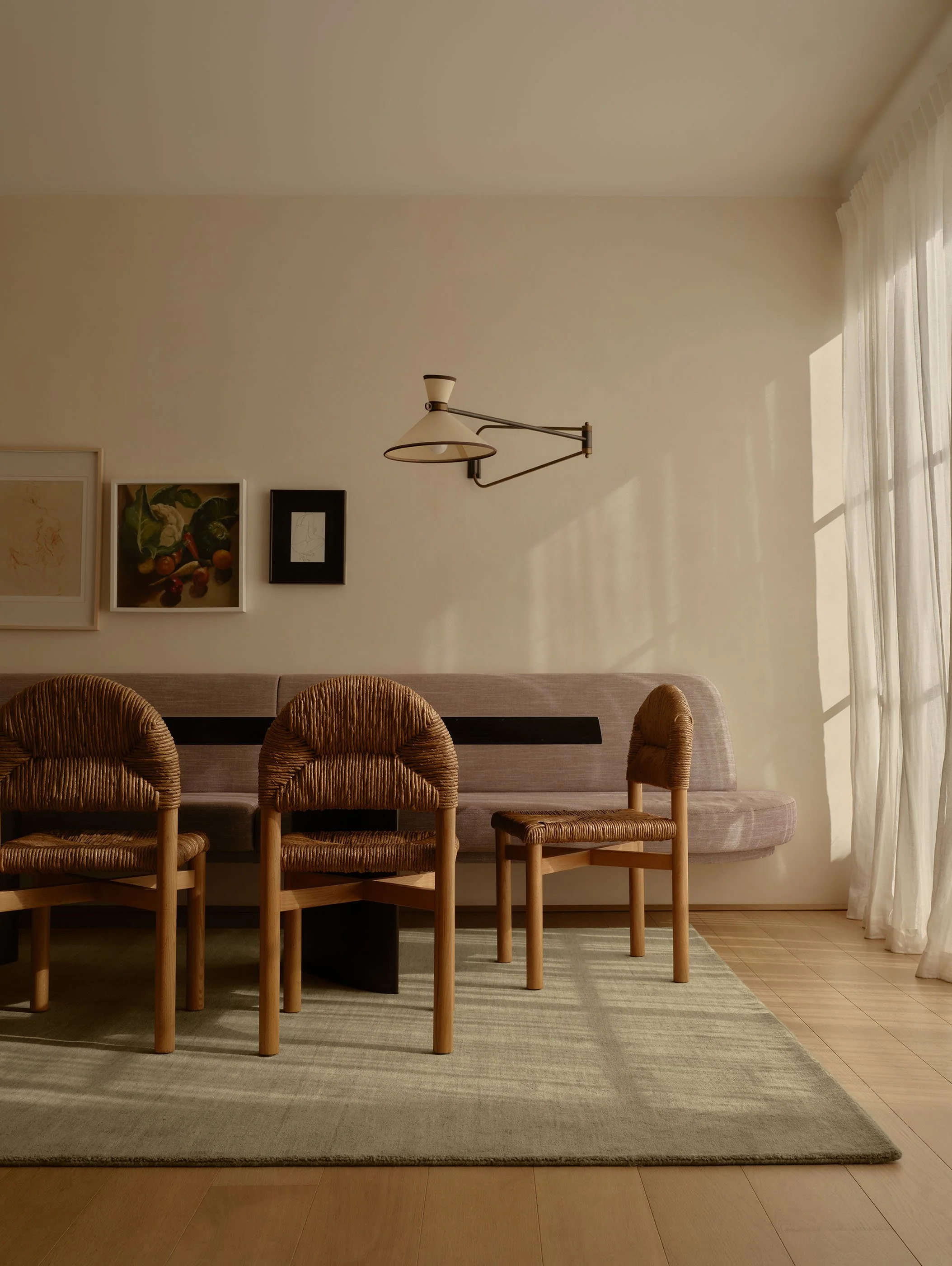 Interior view of a living or dining room with a beige wall, framed artwork, a wall-mounted light fixture, a light gray sofa, wooden chairs with woven seats, a striped rug, and sheer curtains with sunlight streaming in.