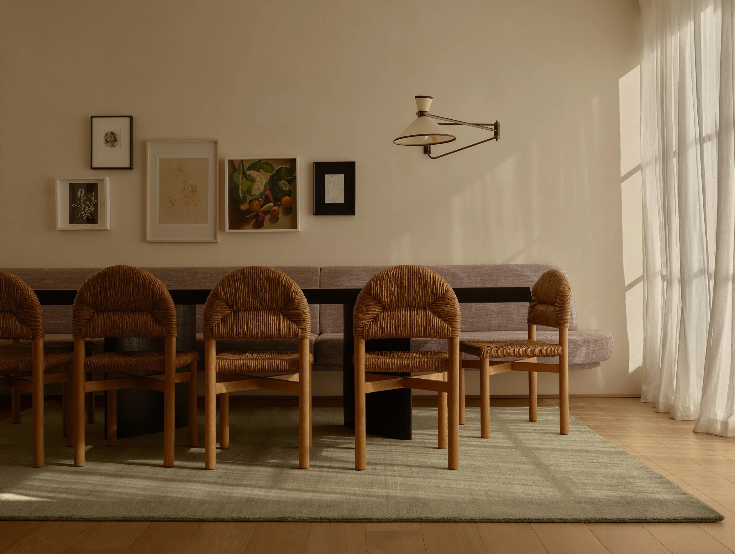 Living room with beige wall, wall art, beige curtains, and a row of six wicker chairs around a dark rectangular table on a rug.