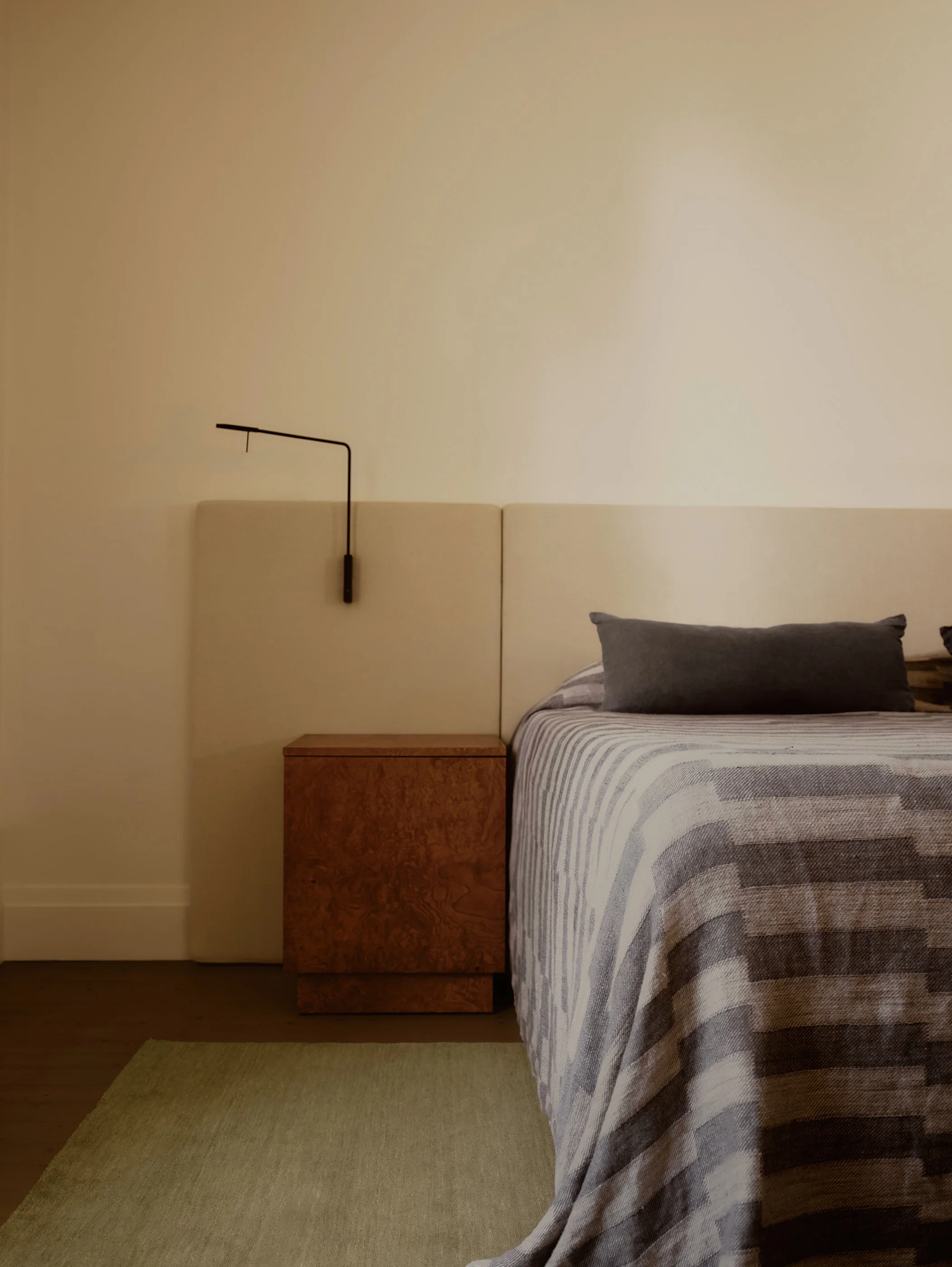 A bedroom with a bed covered in a striped gray and beige blanket, a matching gray pillow, a wooden nightstand, and a black wall-mounted reading lamp, against a plain white wall.