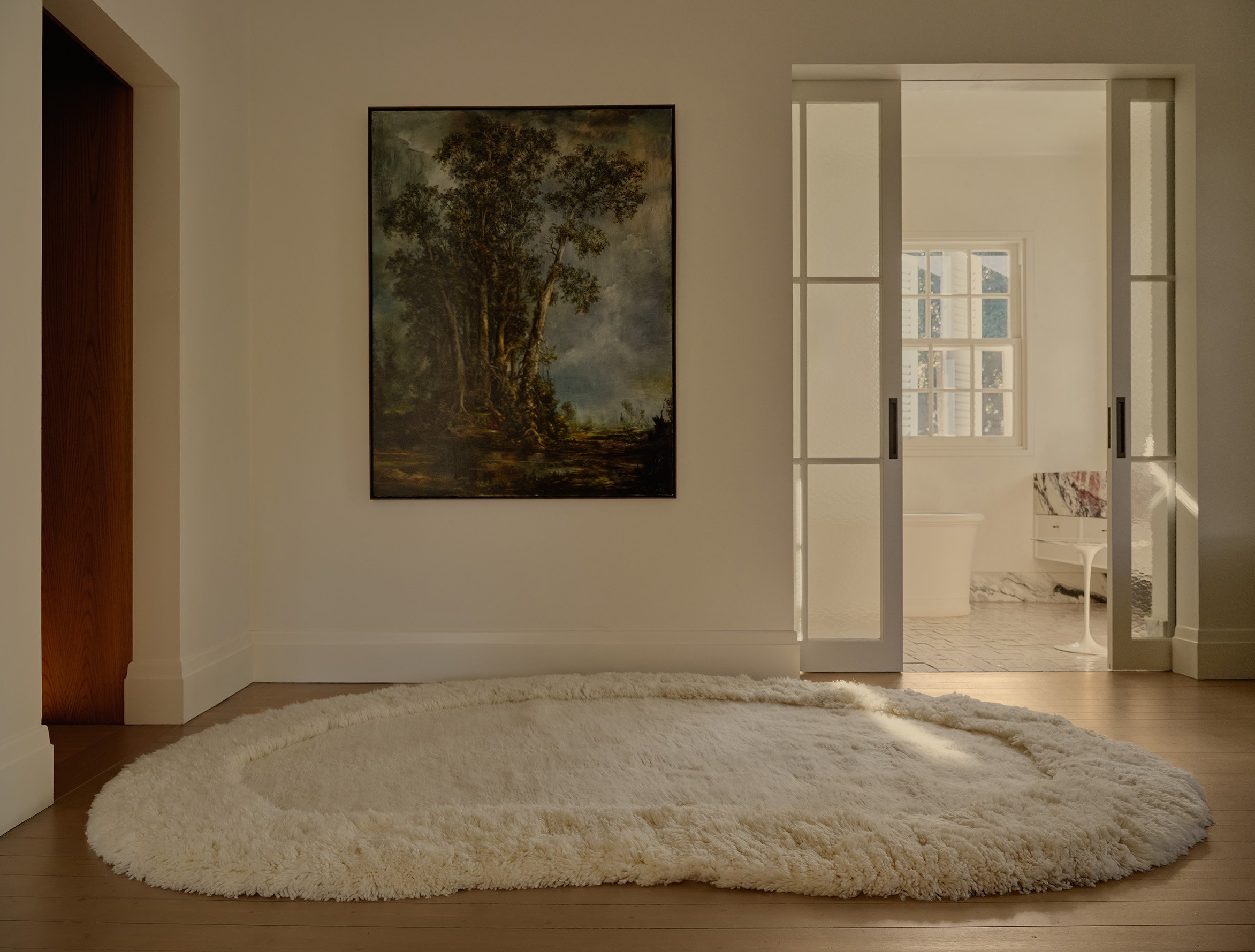 A cozy living room corner with a white shaggy rug on wooden flooring, a wall art depicting a large tree, and a window with white blinds allowing natural light to brighten the space.