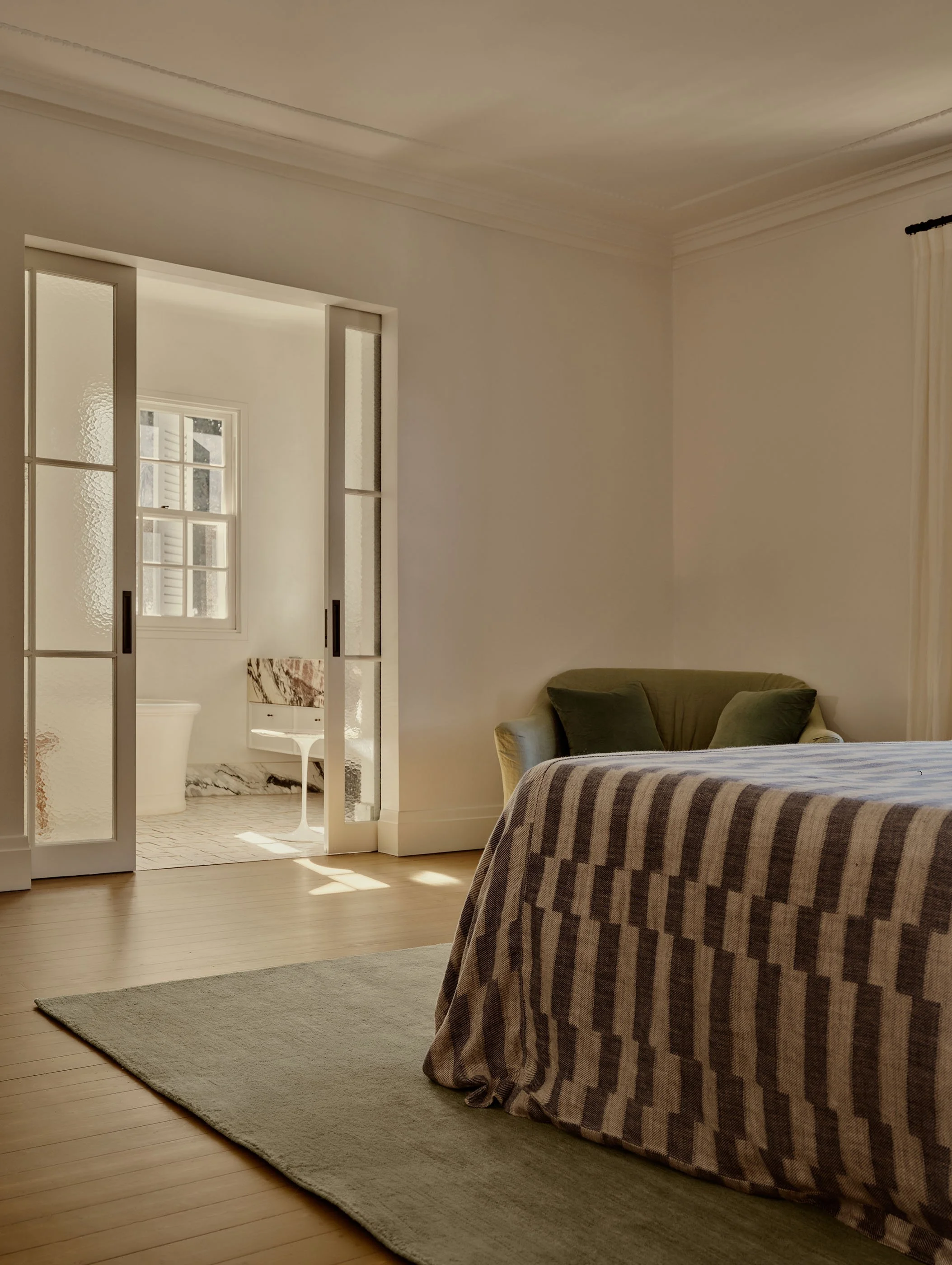 Photo of a cozy room with a bed covered in a plaid blanket, a green armchair, a light-colored rug, and a view into a bathroom through glass-paneled sliding doors, stained glass window, and marble floor in the bathroom.