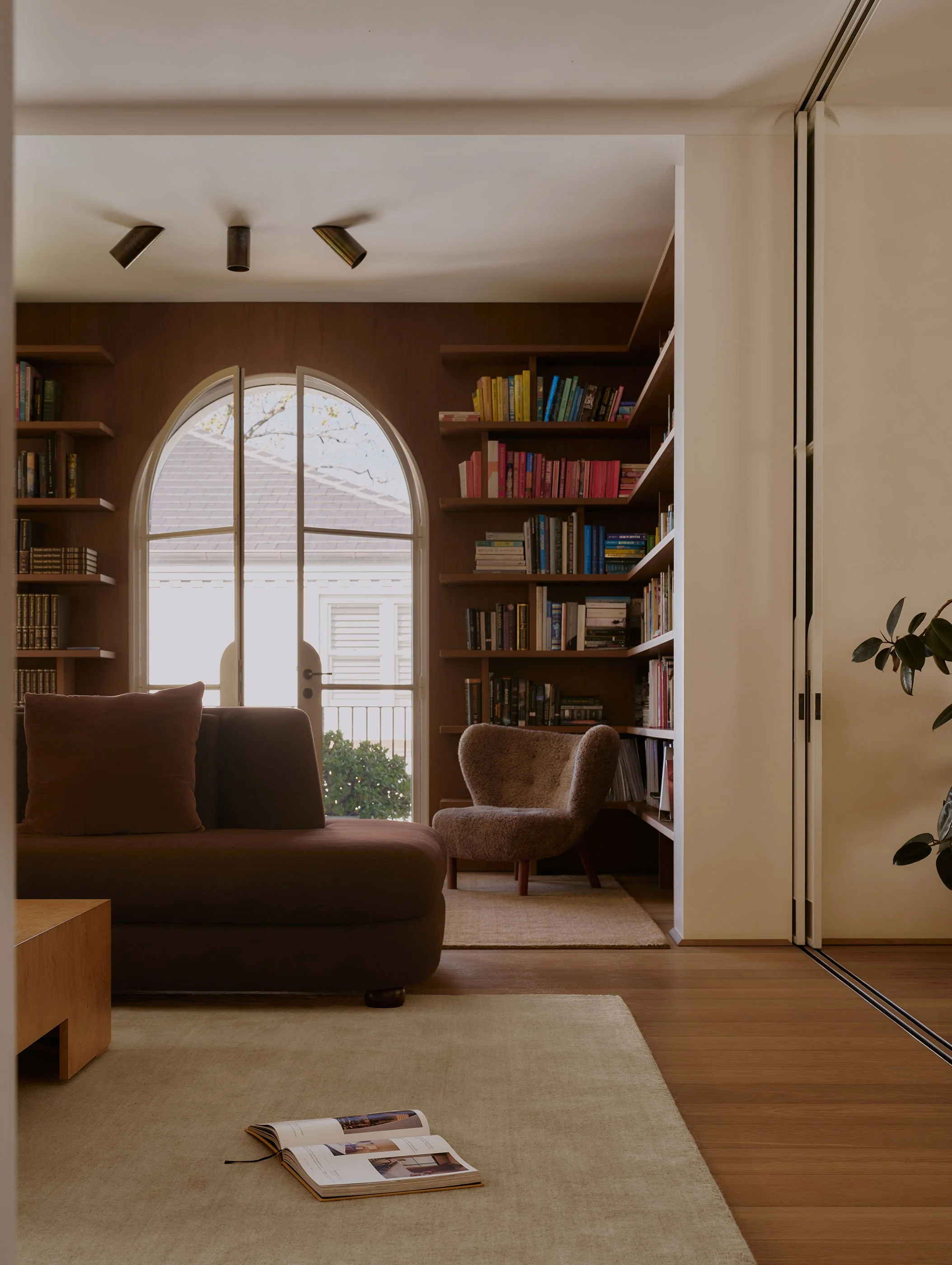 Living room with a brown sofa, open book on beige carpet, large arched window with view of trees, bookshelf filled with colorful books, partly open folding door, potted plant on right