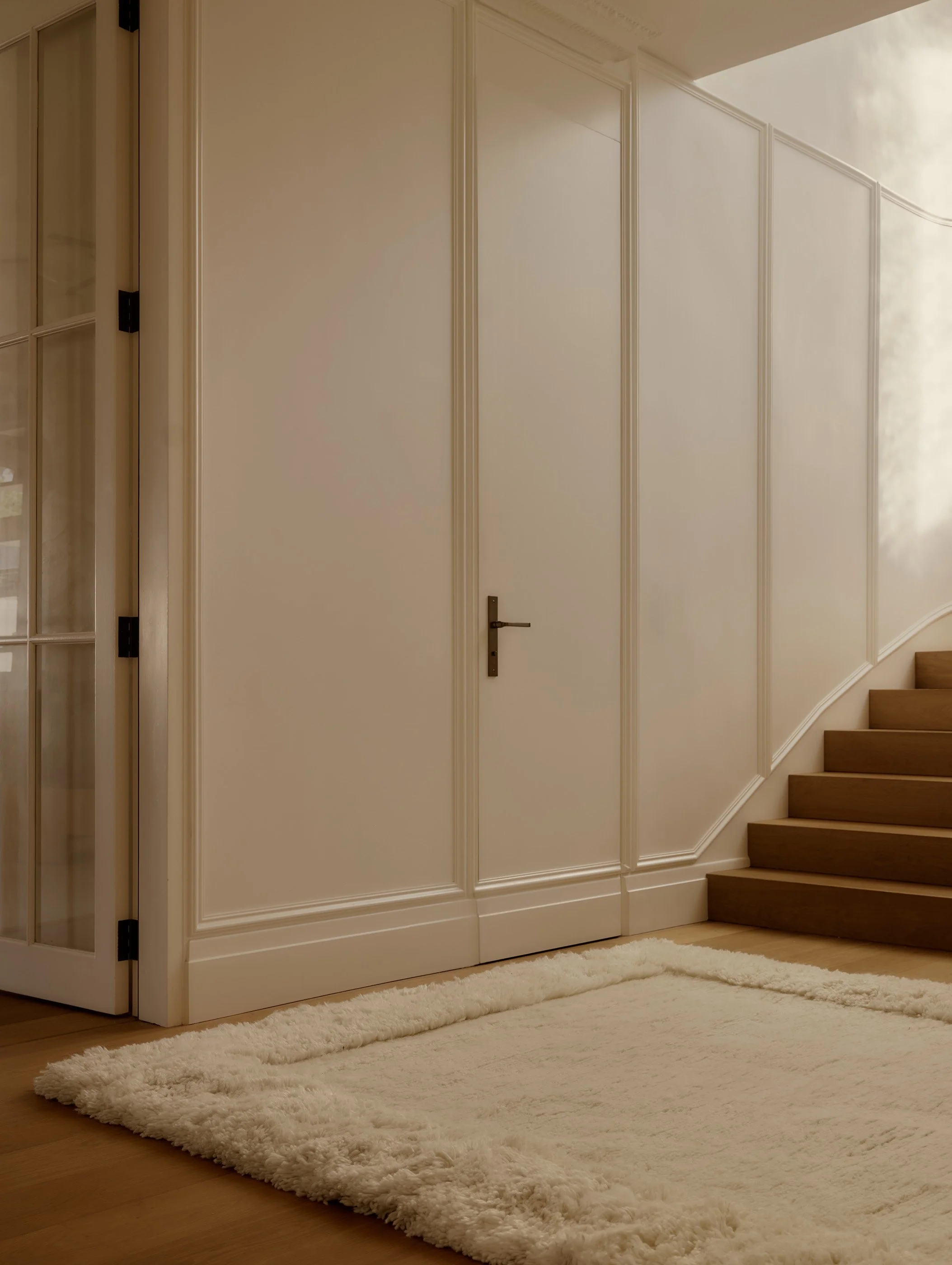 A white door with decorative molding, a black handle, and a floor-length panel, next to a staircase with wooden steps, a white wall, and a cream-colored fluffy rug.