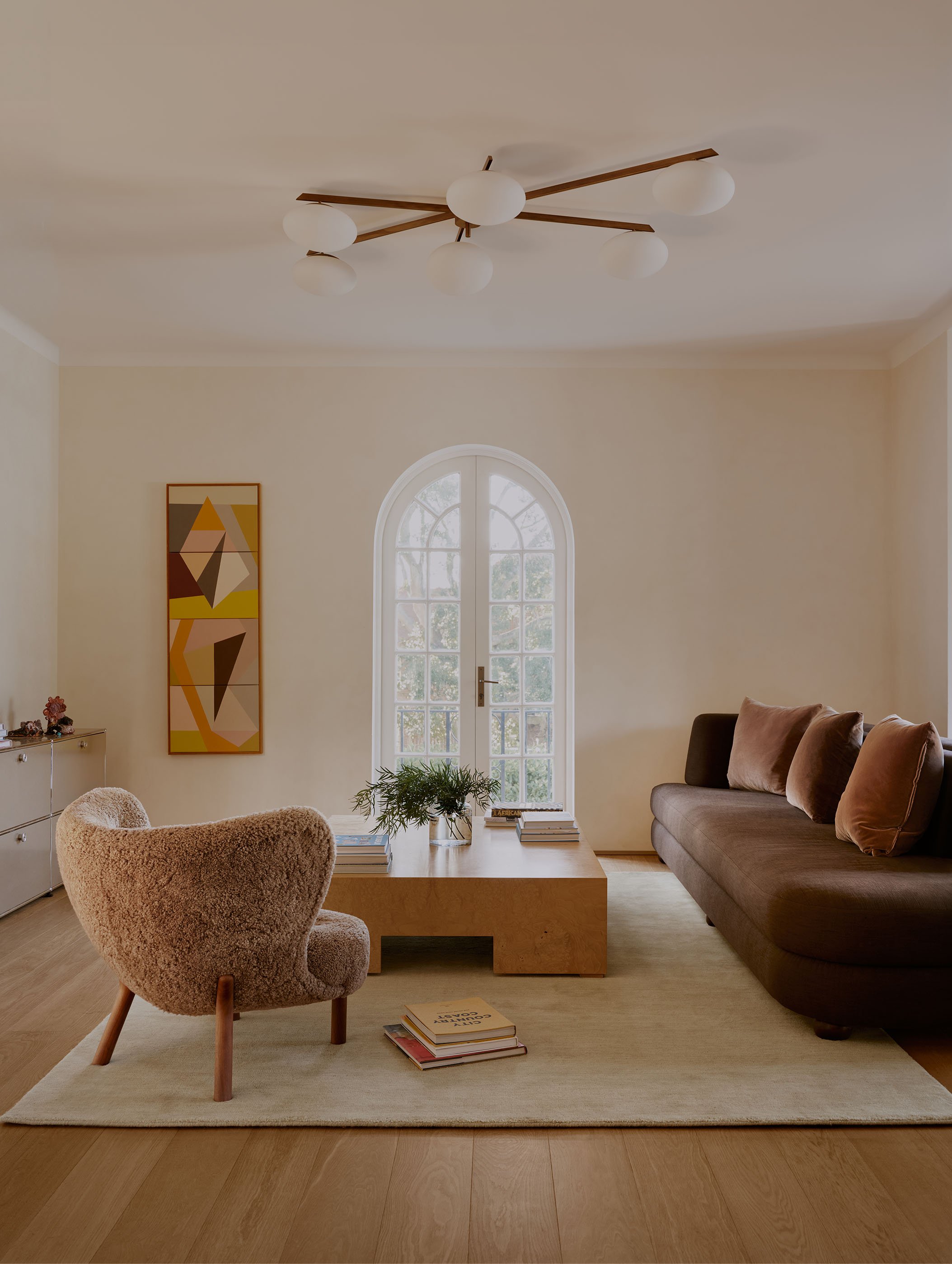 Living room with a modern ceiling light fixture, an arched window, beige walls, abstract artwork, a beige textured armchair, a wooden coffee table with plants and books, a brown sofa with throw pillows, and a small white dresser with decorative items