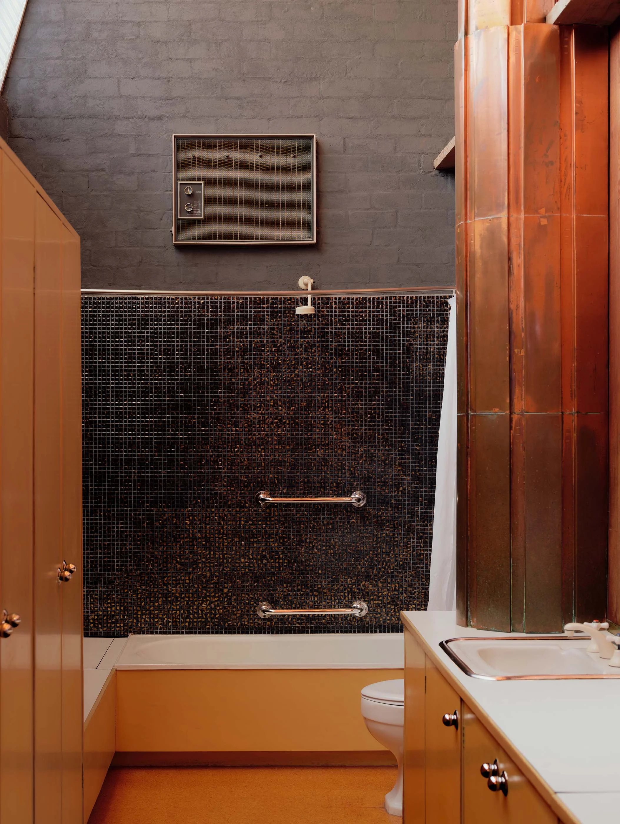 This is a small bathroom with a shower area that has dark mosaic tiles, a wall-mounted heater, a white toilet, and a beige wooden vanity with a sink. The walls are a mix of dark and warm tones, with a small section of window visible in the upper left