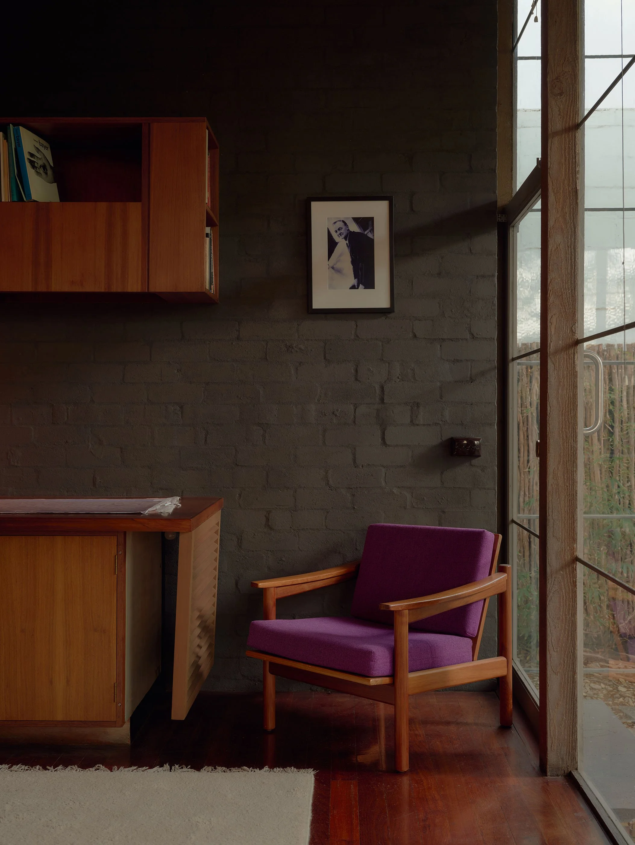 A cozy indoor corner with a purple armchair, a wooden bookshelf with books, a framed black-and-white photo on a brick wall, a window with a wooden frame, and a beige rug on a wooden floor.
