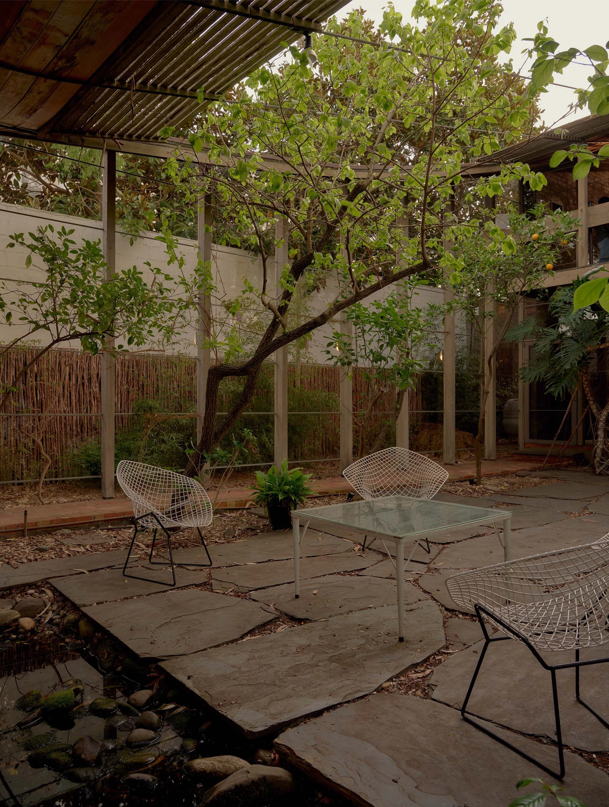 A backyard patio with a small pond, surrounded by rocks, and a tree with green leaves. There are white wire chairs and a glass-top table on the stone patio, with a wooden balcony above. The scene looks calm and inviting.