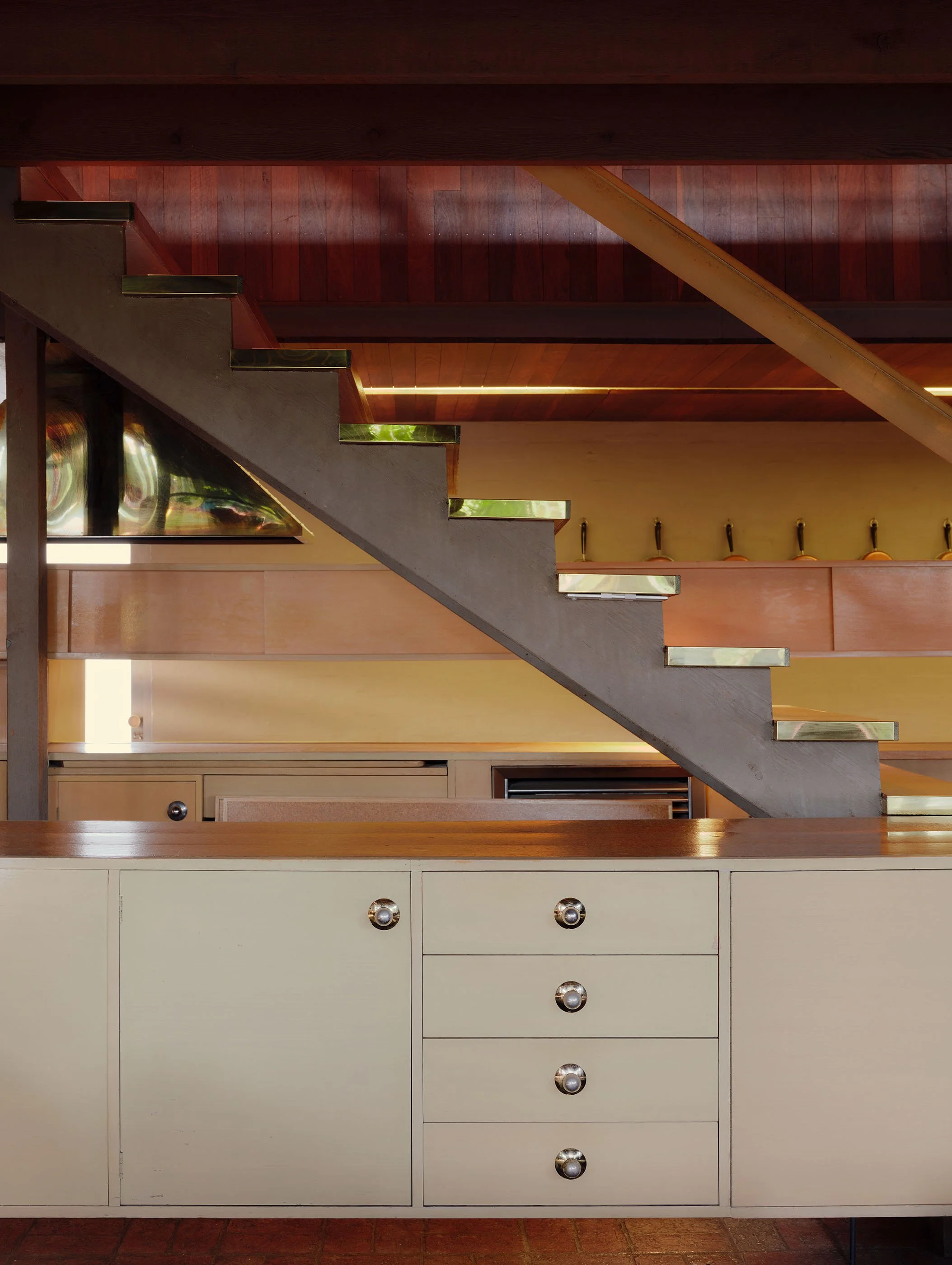 Interior view of a modern kitchen with a staircase. The lower section features a cream-colored cabinet with round metallic handles. The staircase has wooden steps with metallic accents on the edges. Behind the staircase, there is a wooden-paneled wal