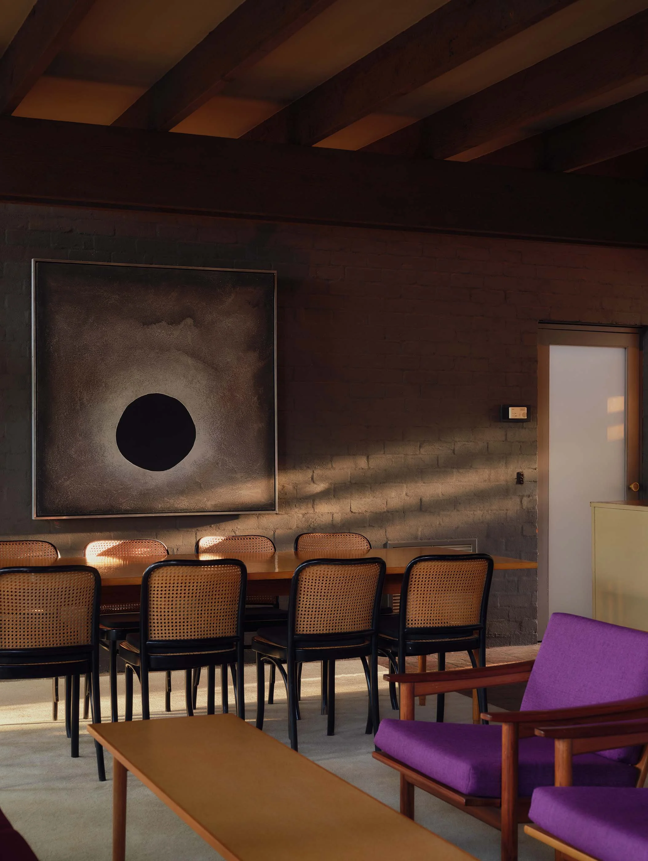 Interior of a room with a wooden table, purple armchairs, a long dining table with woven chairs, a brick wall, a piece of abstract art with a black circle on a textured background, a window with frosted glass, and a wall-mounted thermostat.