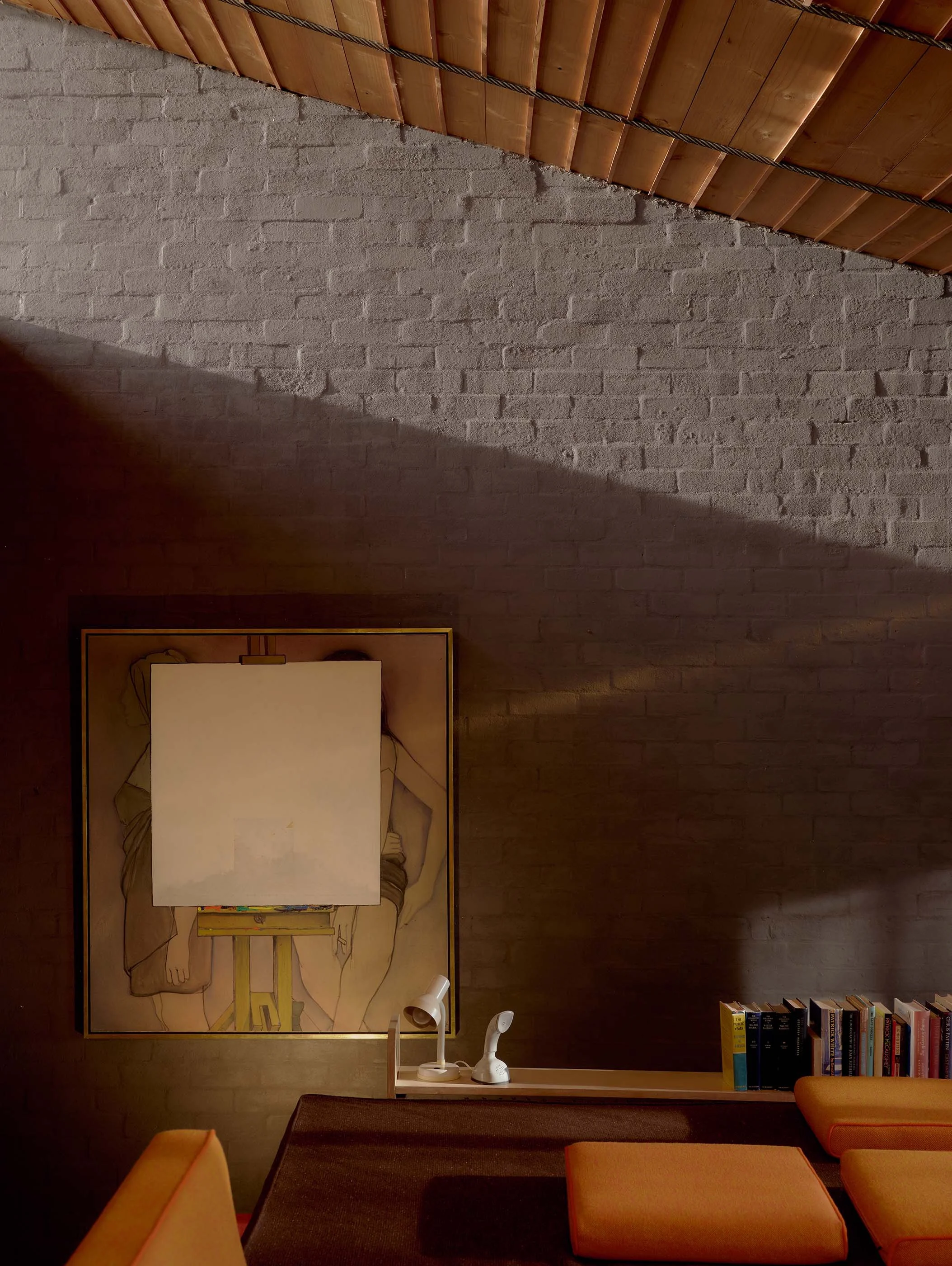 Living room with a large framed artwork of a woman lying on her side, a white lamp, a set of books on a shelf, and a beige couch with orange cushions. The room features a brick wall and a wooden ceiling with exposed beams.