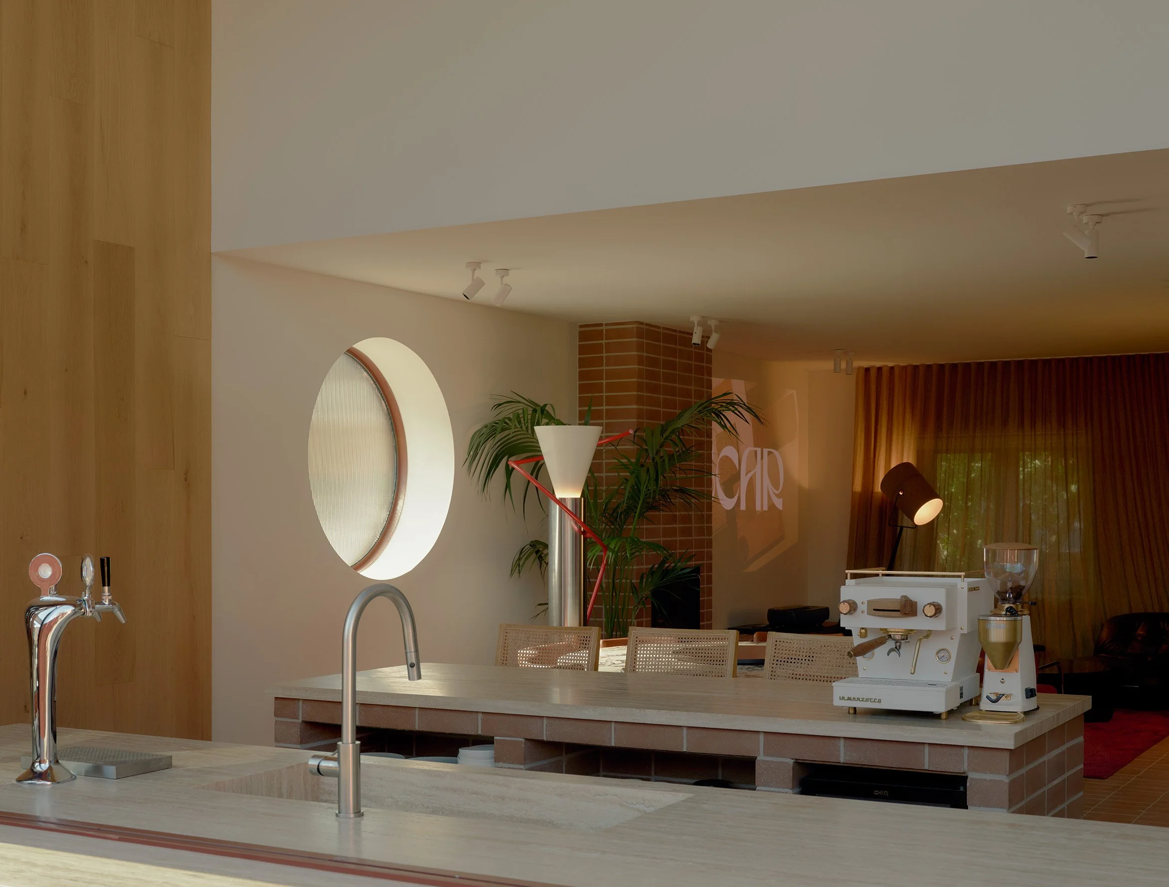 Modern kitchen with light-colored wooden countertops and cabinets, a circular window, potted plants, and vintage-style kitchen appliances, including a pasta machine on the counter.