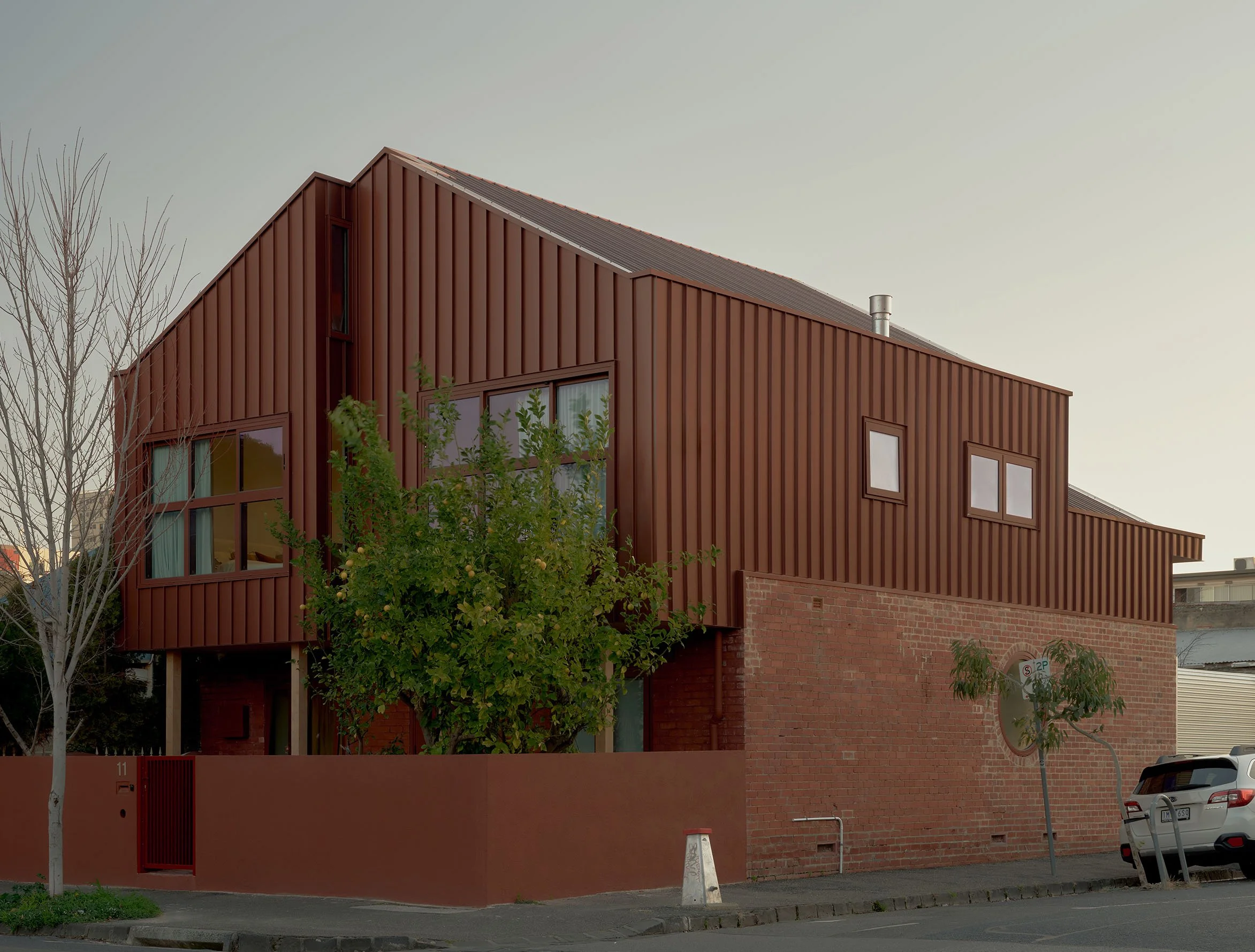 Modern building with red metal siding, multiple windows, and a tree in front, parked cars, and a brick wall at the base.