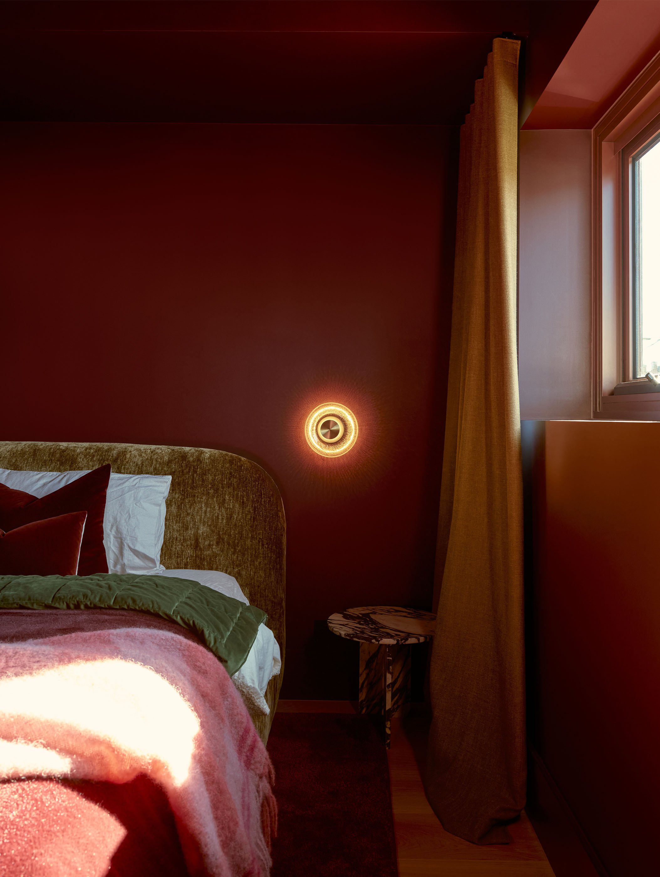 Interior view of a bedroom with a bed, pillows, a pink blanket, a dark red wall, a round wall light, a small side table with a black and white marble pattern, a window with yellow curtains, and natural light coming in.