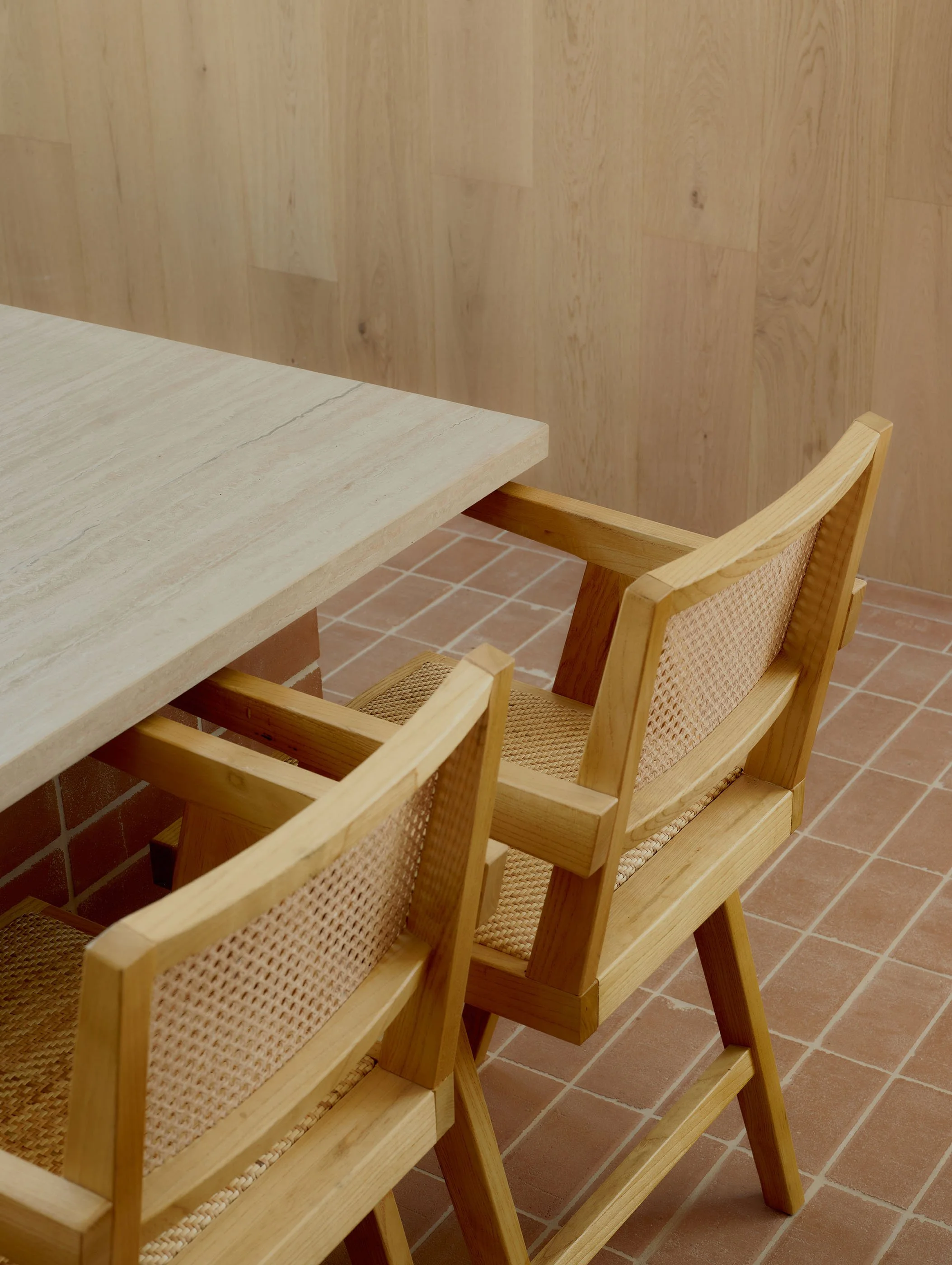 The image shows a wooden dining chair with a woven rattan backrest next to a wood table with a light-colored stone or marble top, set against a wooden wall and terracotta-tiled floor.
