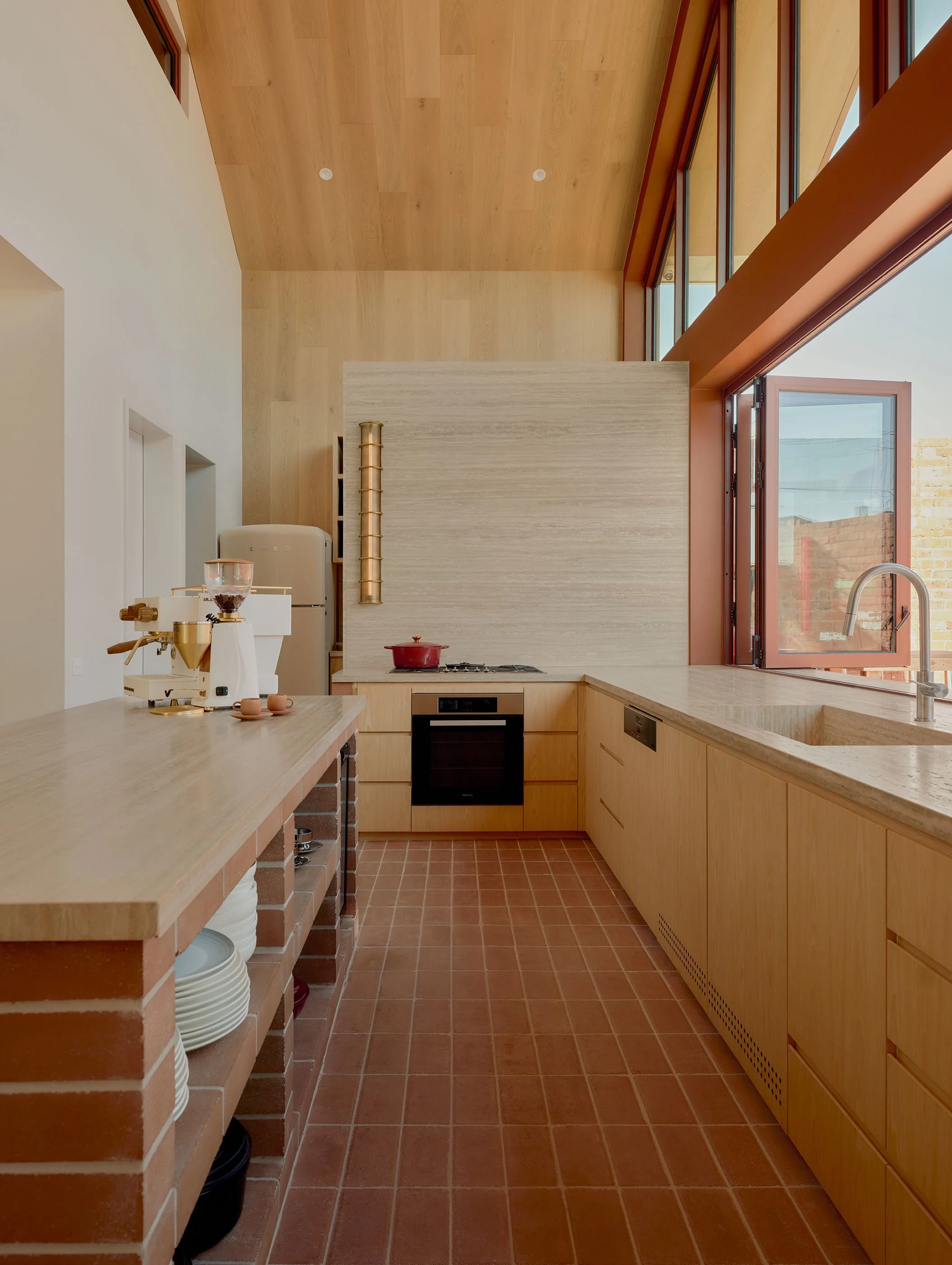 Modern kitchen with light wood cabinets, terracotta tiled floor, and large windows with orange frames letting in natural light.