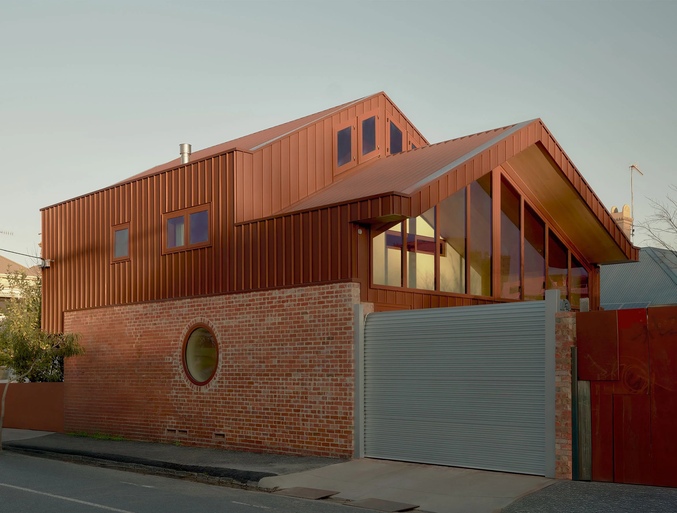 Modern building with red metal siding, large glass windows, and a brick base, located on a city street with a gray metal gate and surrounding trees.