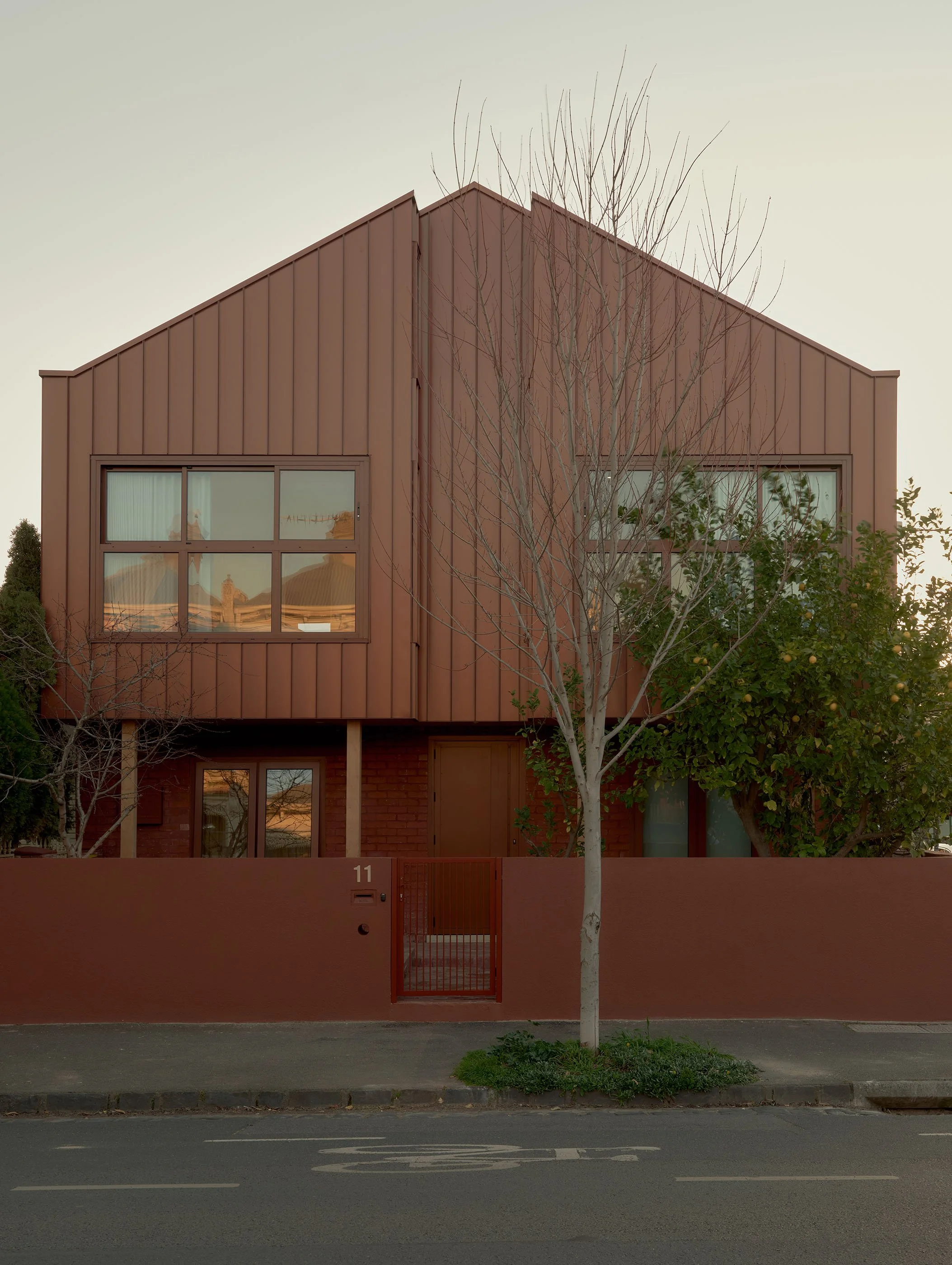 A modern two-story house painted in reddish-brown with large rectangular windows, a flat roof, and a matching reddish-brown wall and gate. A leafless tree and some greenery are in front of the house, and there is a sidewalk and street with lane marki
