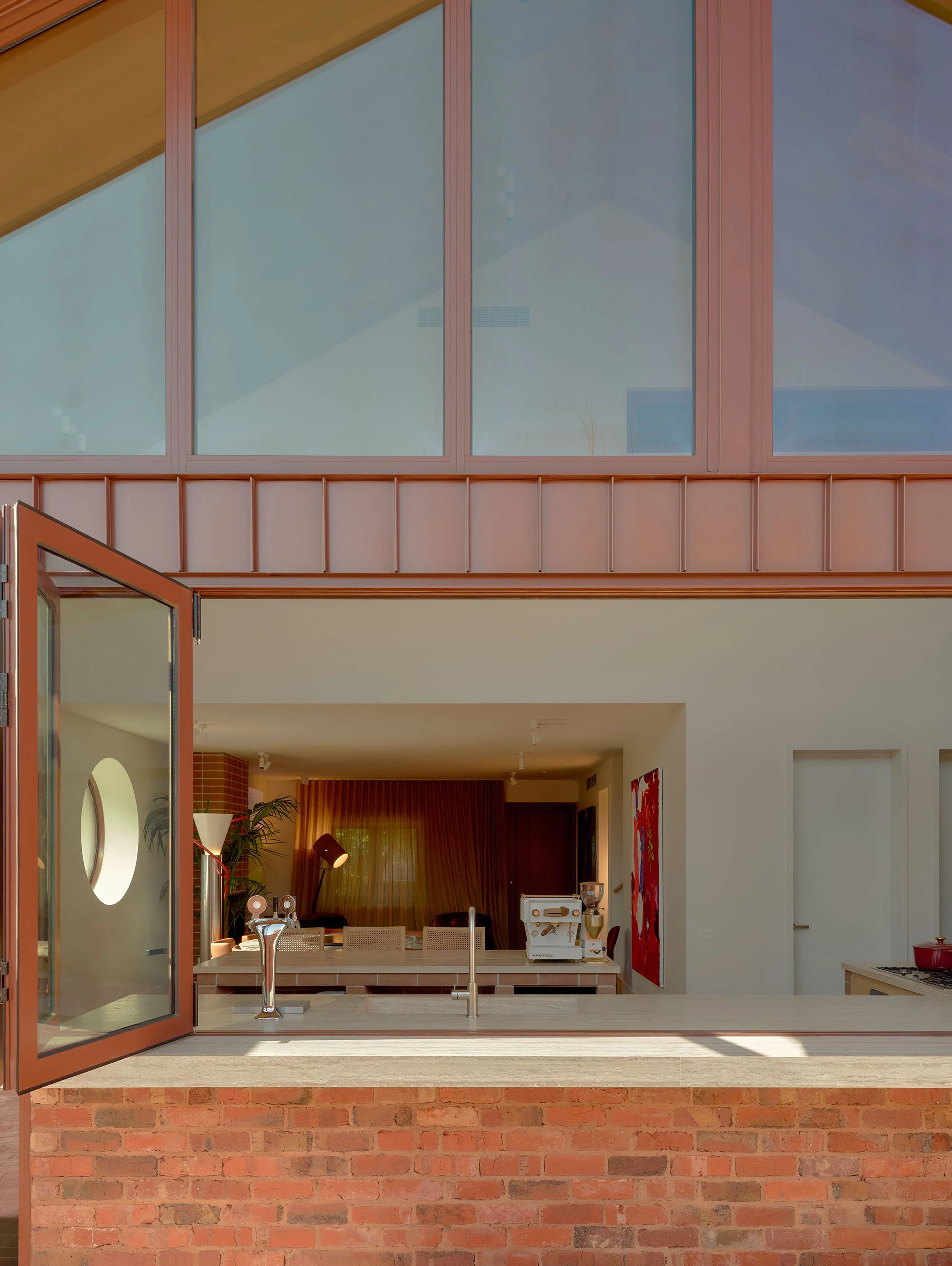 View of a modern kitchen with an open window looking into a room with living space, including a dining area and art on the walls.