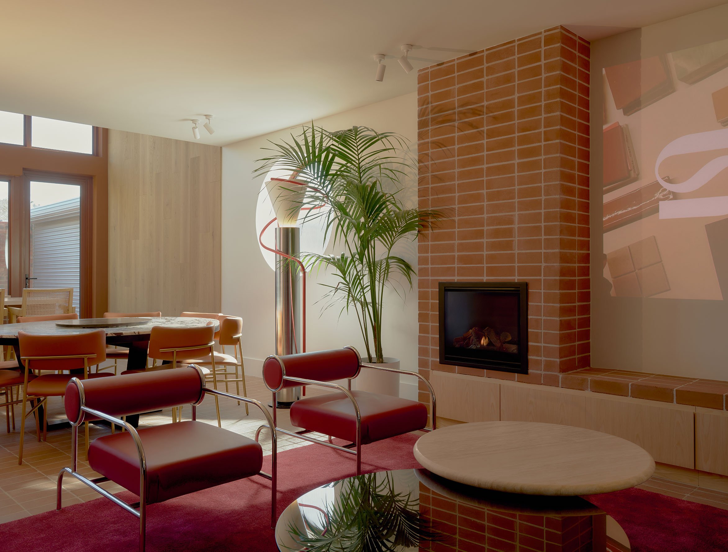 Interior living room with red chairs, a wooden table, a brick fireplace, a potted plant, window, and wall art.