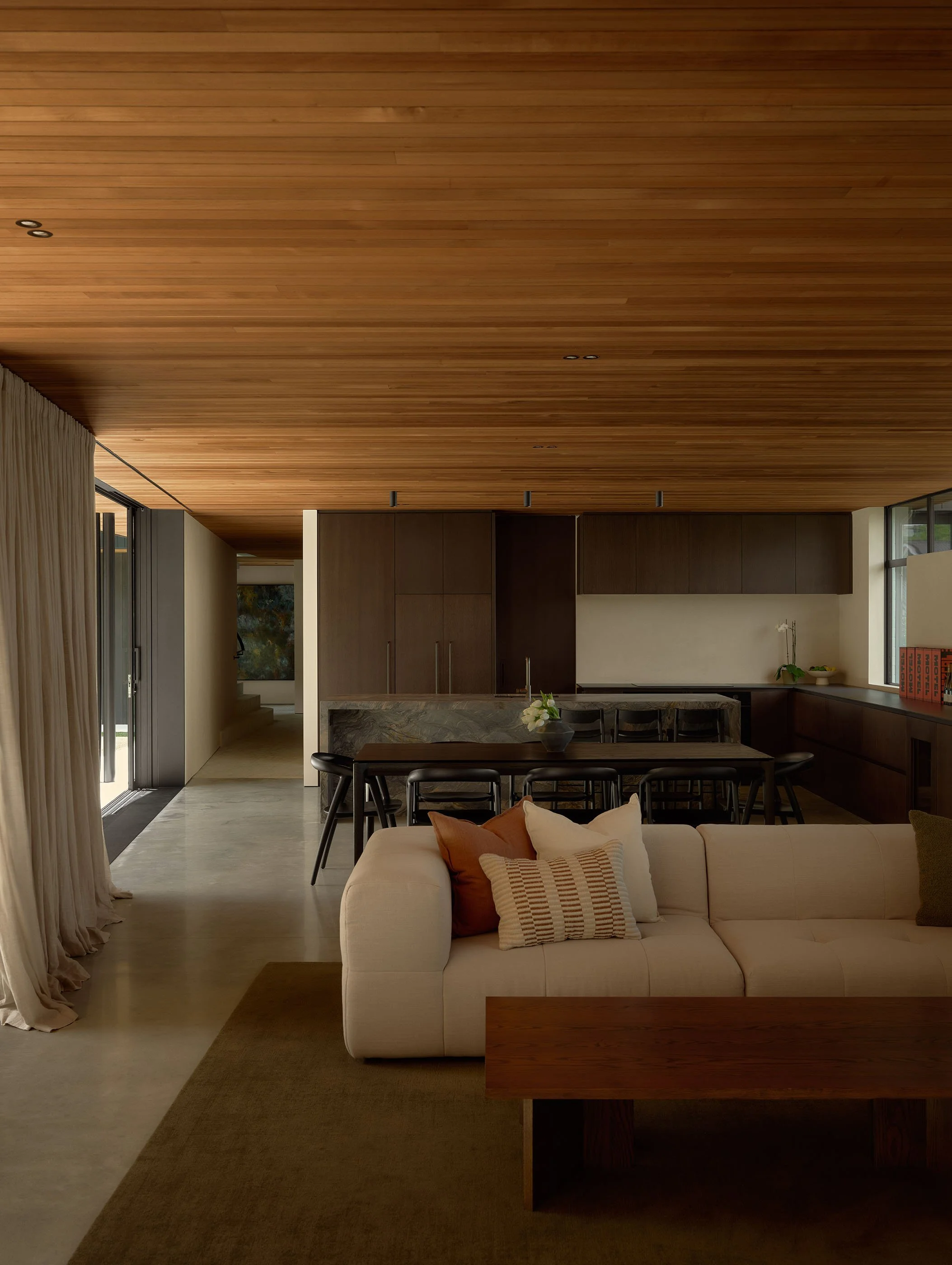 A modern open-concept living space with a high wooden ceiling, a white sofa with pillows, a kitchen area with dark wood cabinets, a dining table with chairs, and large windows allowing natural light.
