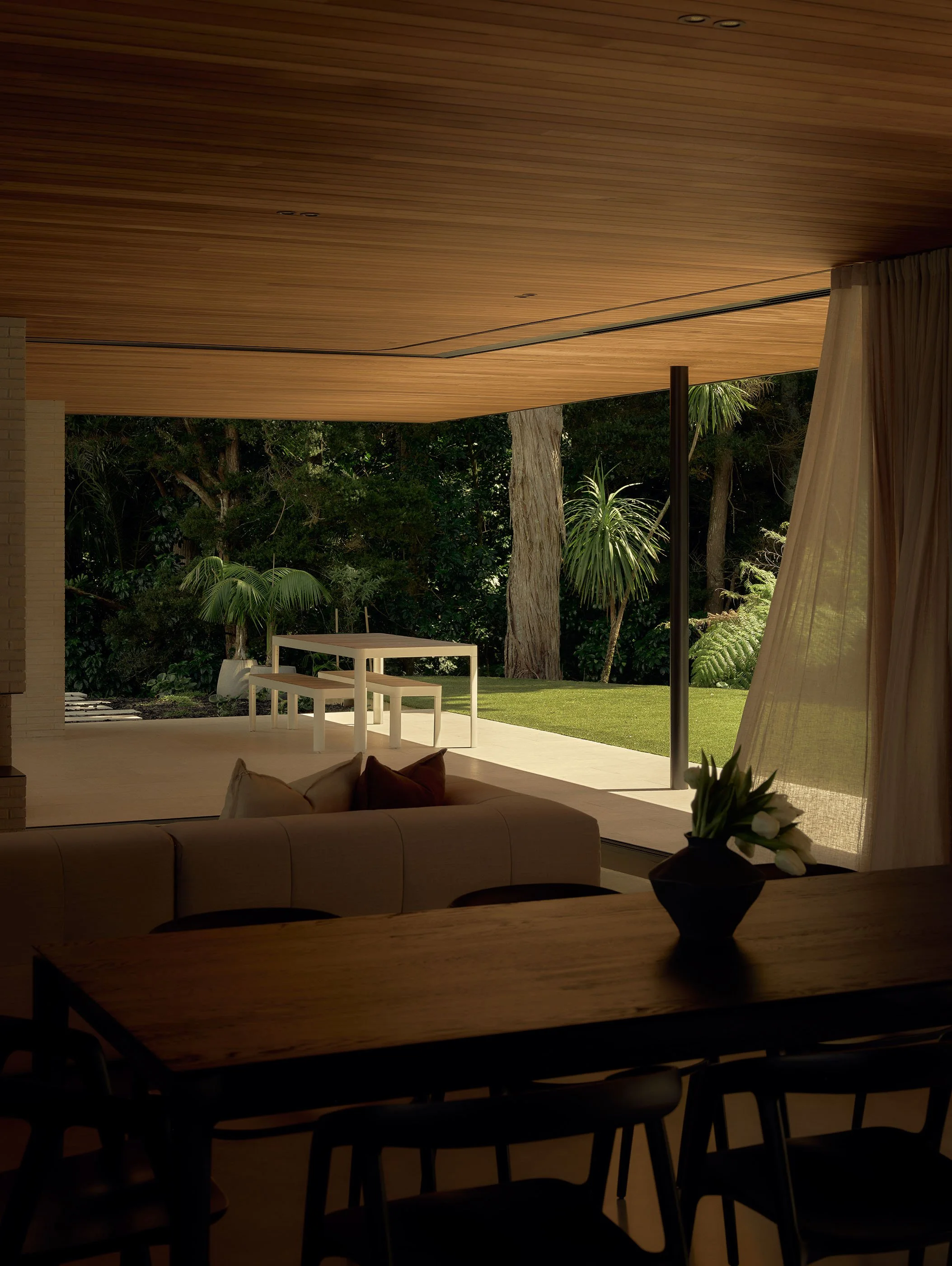 Indoor view of a modern living space with wooden ceiling, large windows open to a garden with trees and plants, light-colored curtains, a white table with bench, and a sofa with cushions.