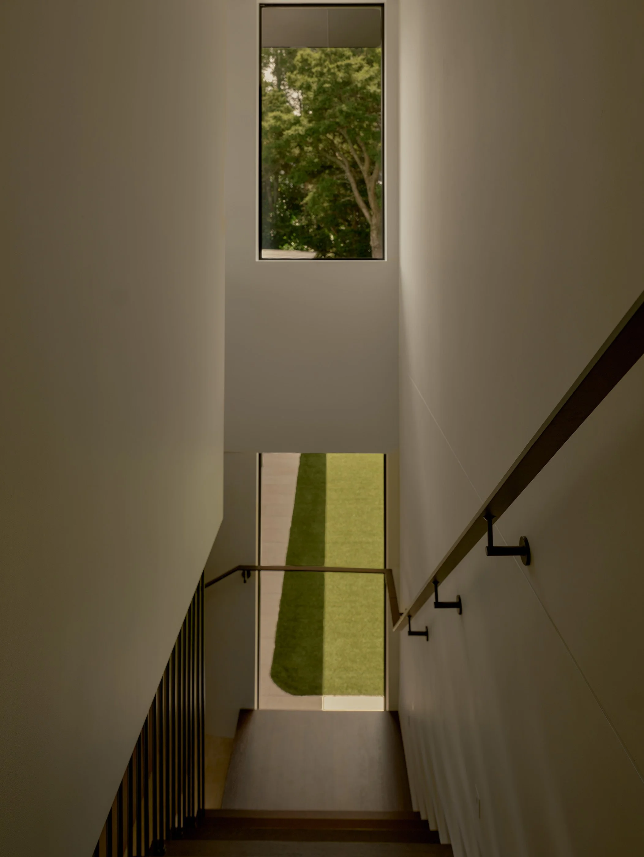 View from the bottom of a staircase looking up through a narrow opening with a window, showing green trees outside.
