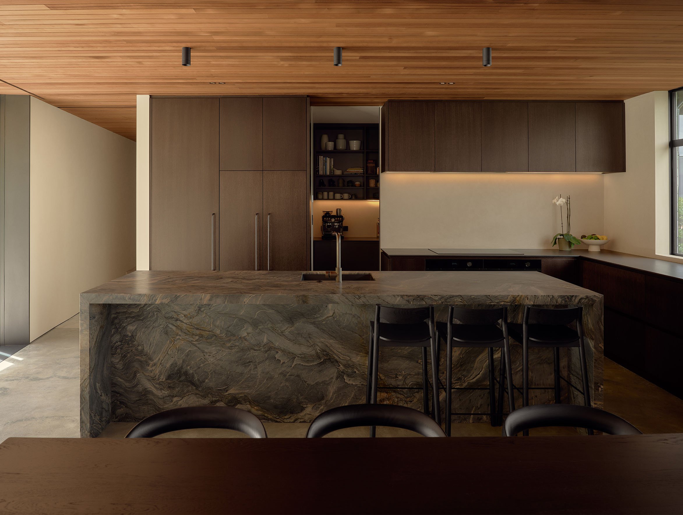 Modern kitchen with a large marble island, wooden cabinetry, and a coffee machine in a nook with dishware, illuminated by natural light through a window.