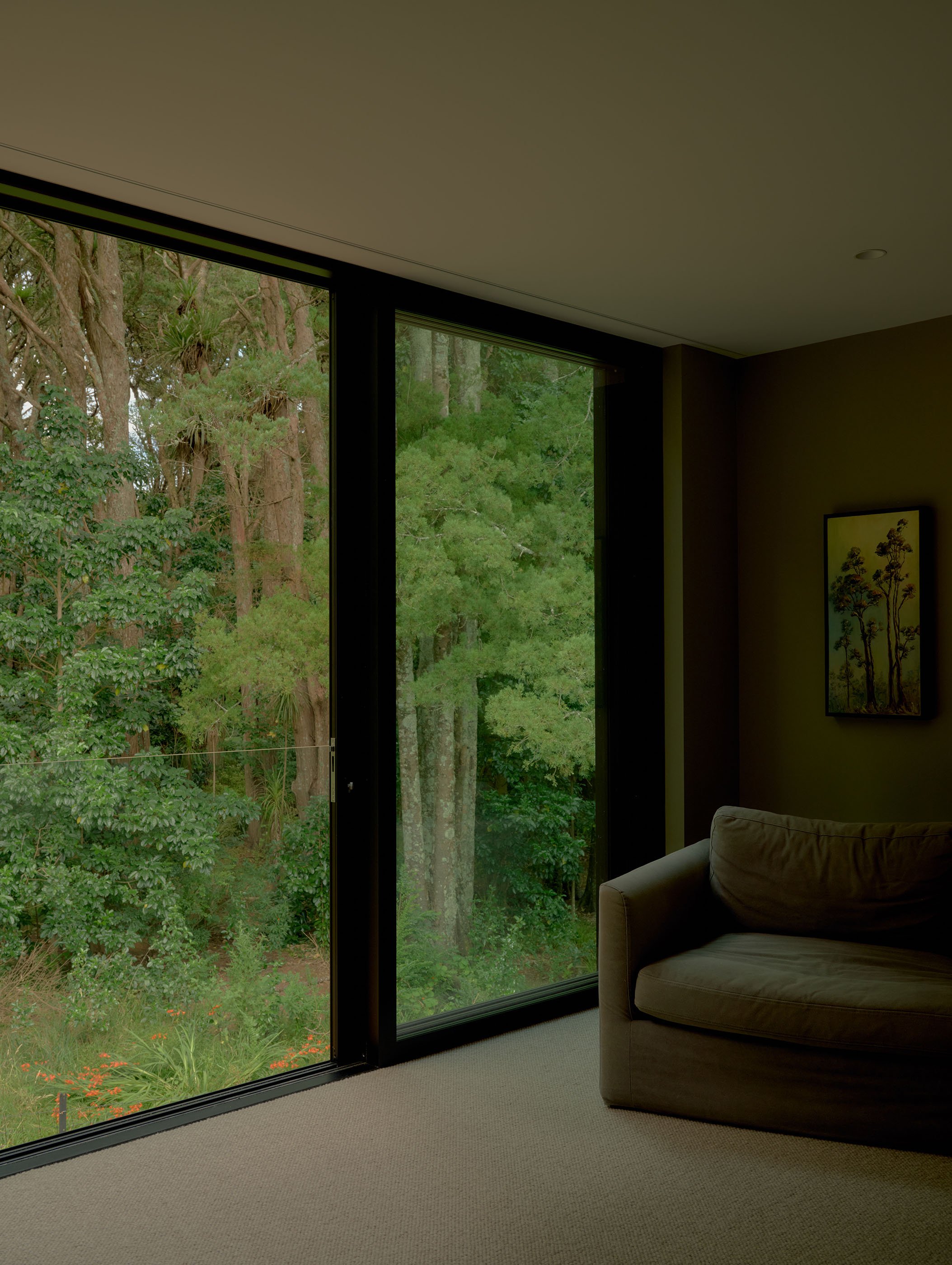 Living room with large floor-to-ceiling glass windows showing green trees outside, beige carpet, and a brown armchair.