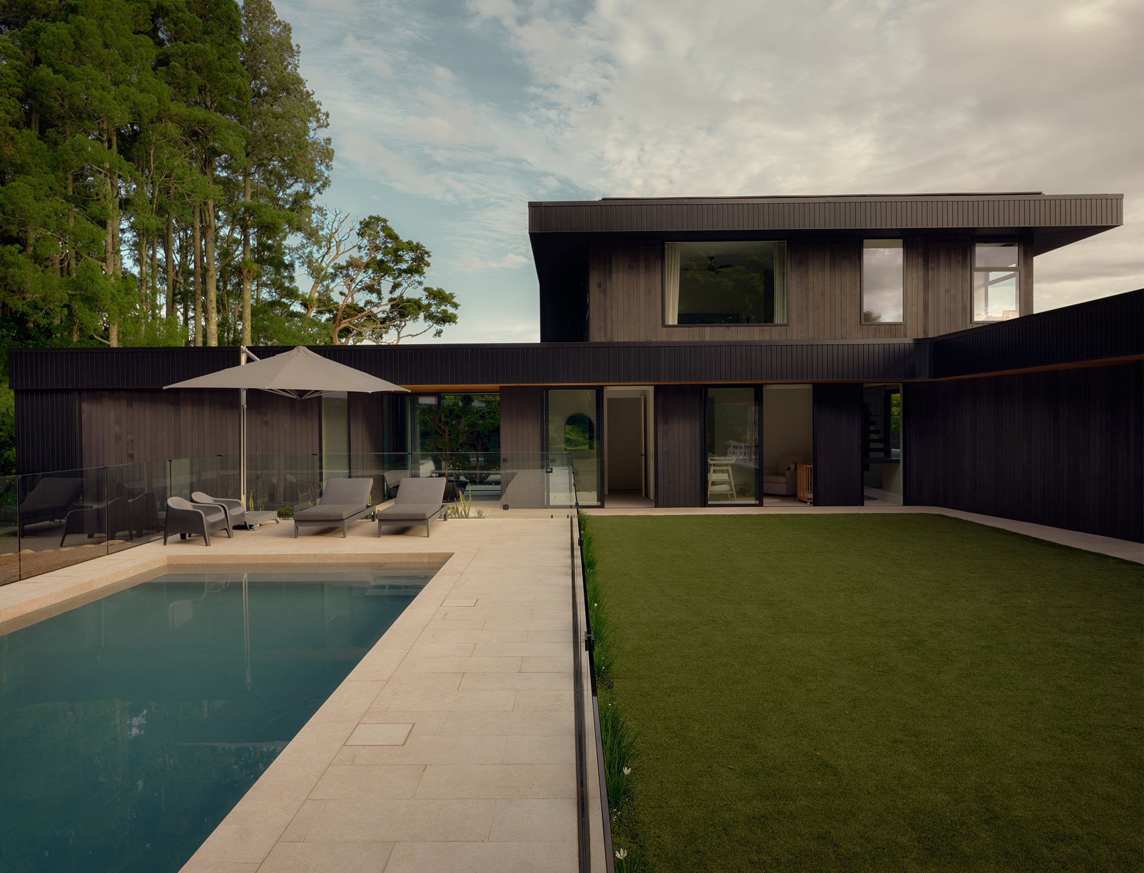 Modern black house with large windows, surrounded by green trees, with a backyard swimming pool and patio area.