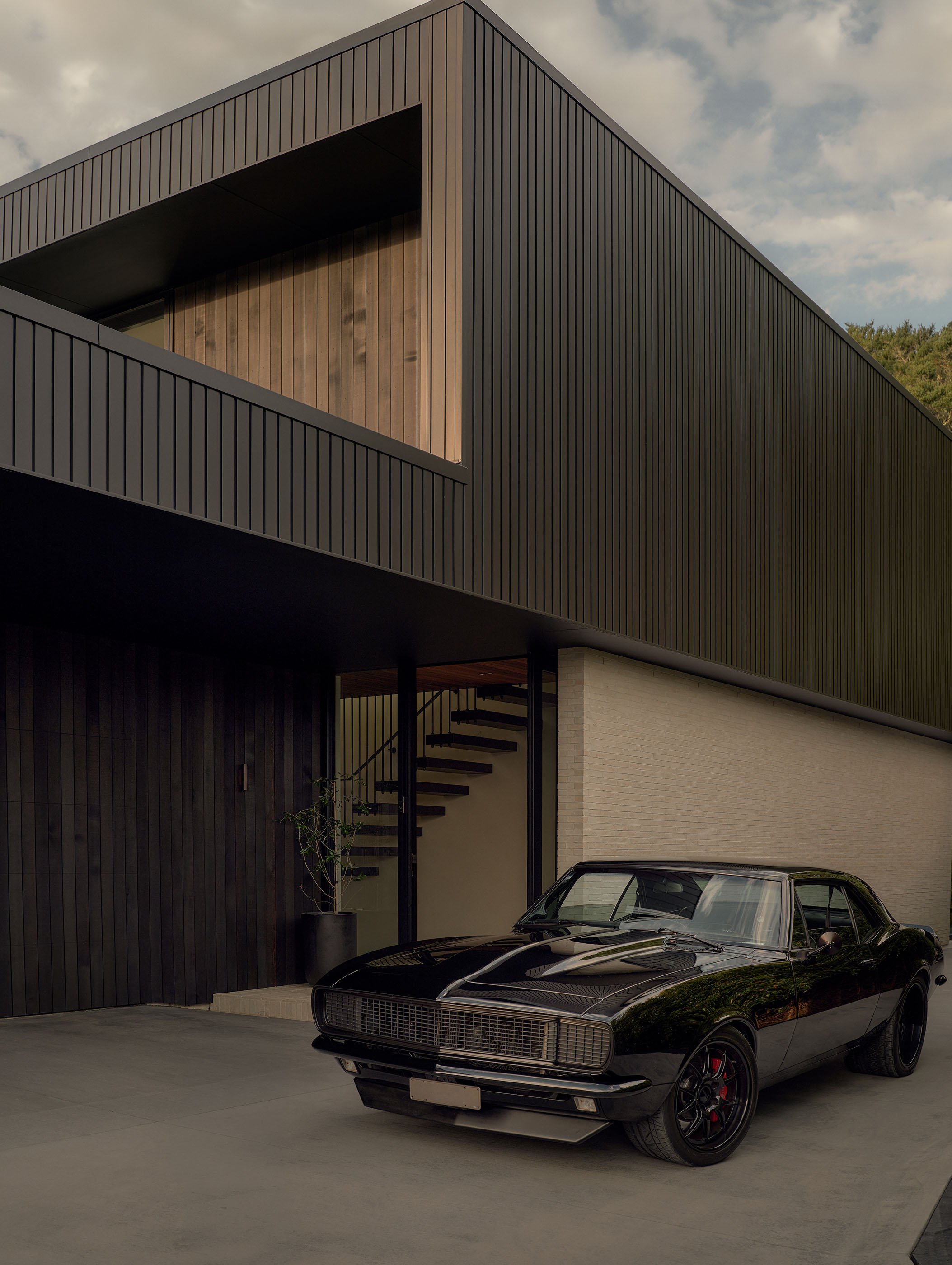 A black vintage car parked in front of a modern building with black vertical siding and a balcony with a wooden interior.