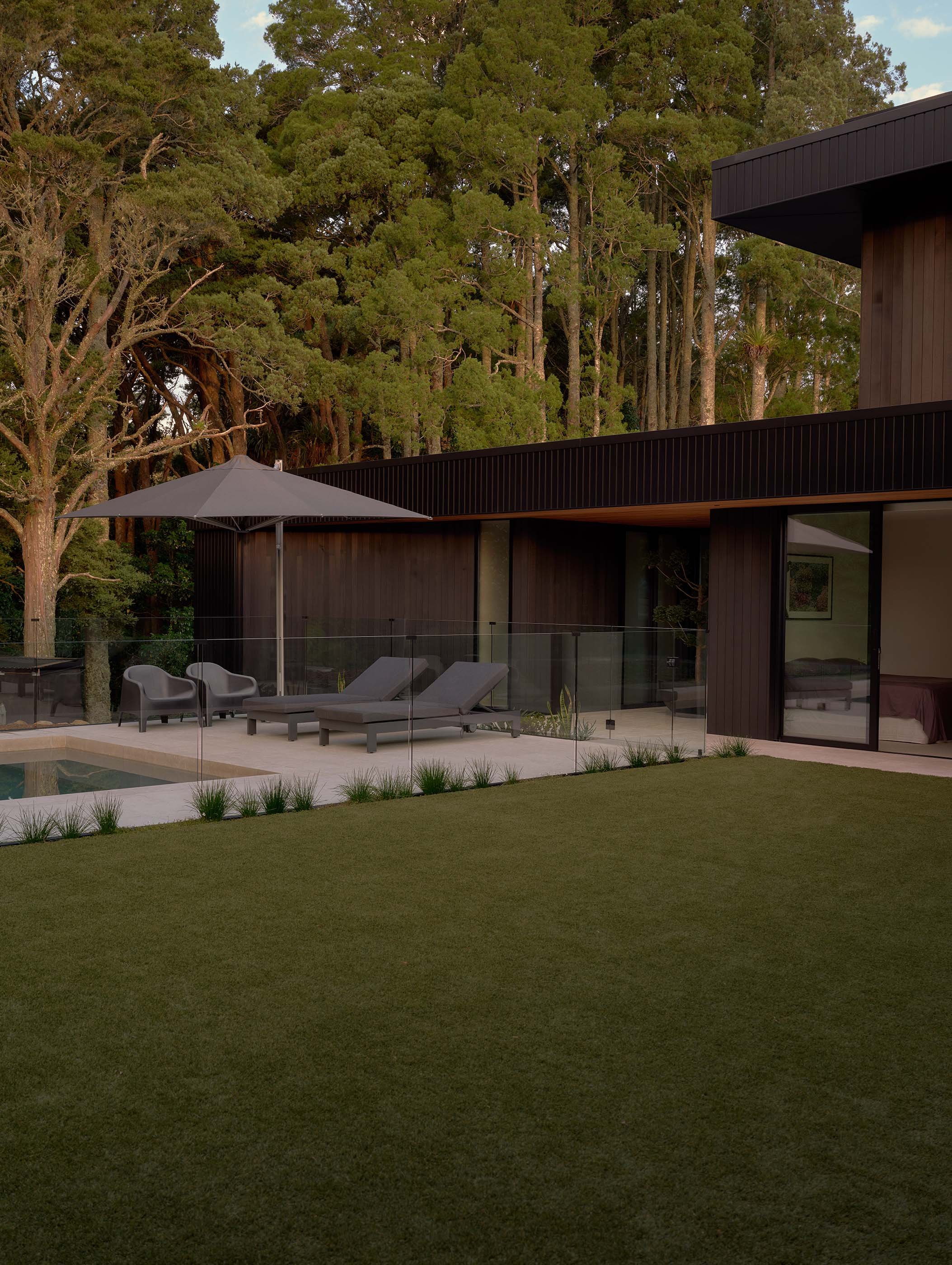 Modern black house with large glass windows, outdoor lounge chairs, patio umbrella, and lush green lawn, with a backdrop of tall trees.