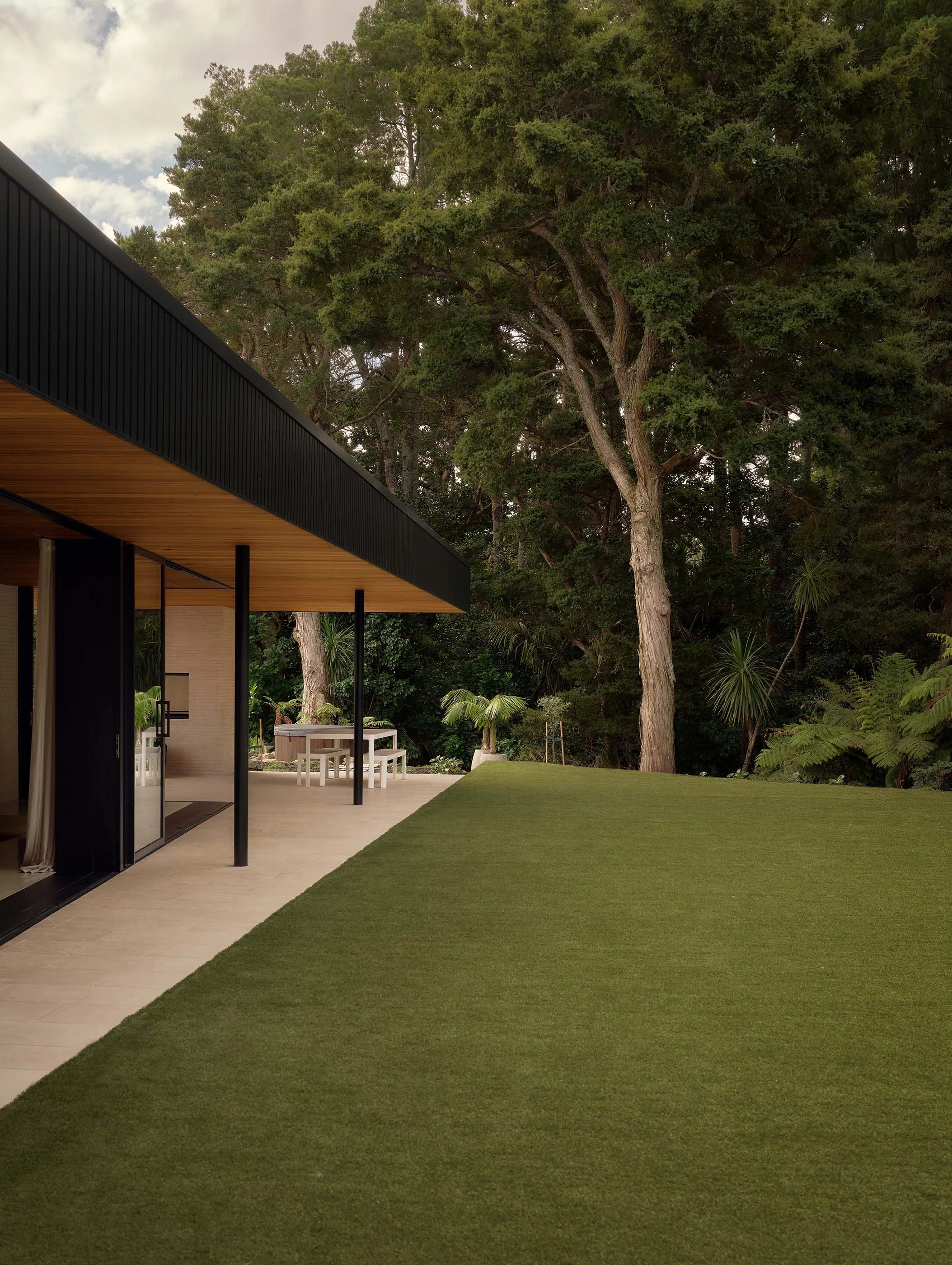 Modern house with a black and wooden overhang, surrounded by lush greenery and large trees.