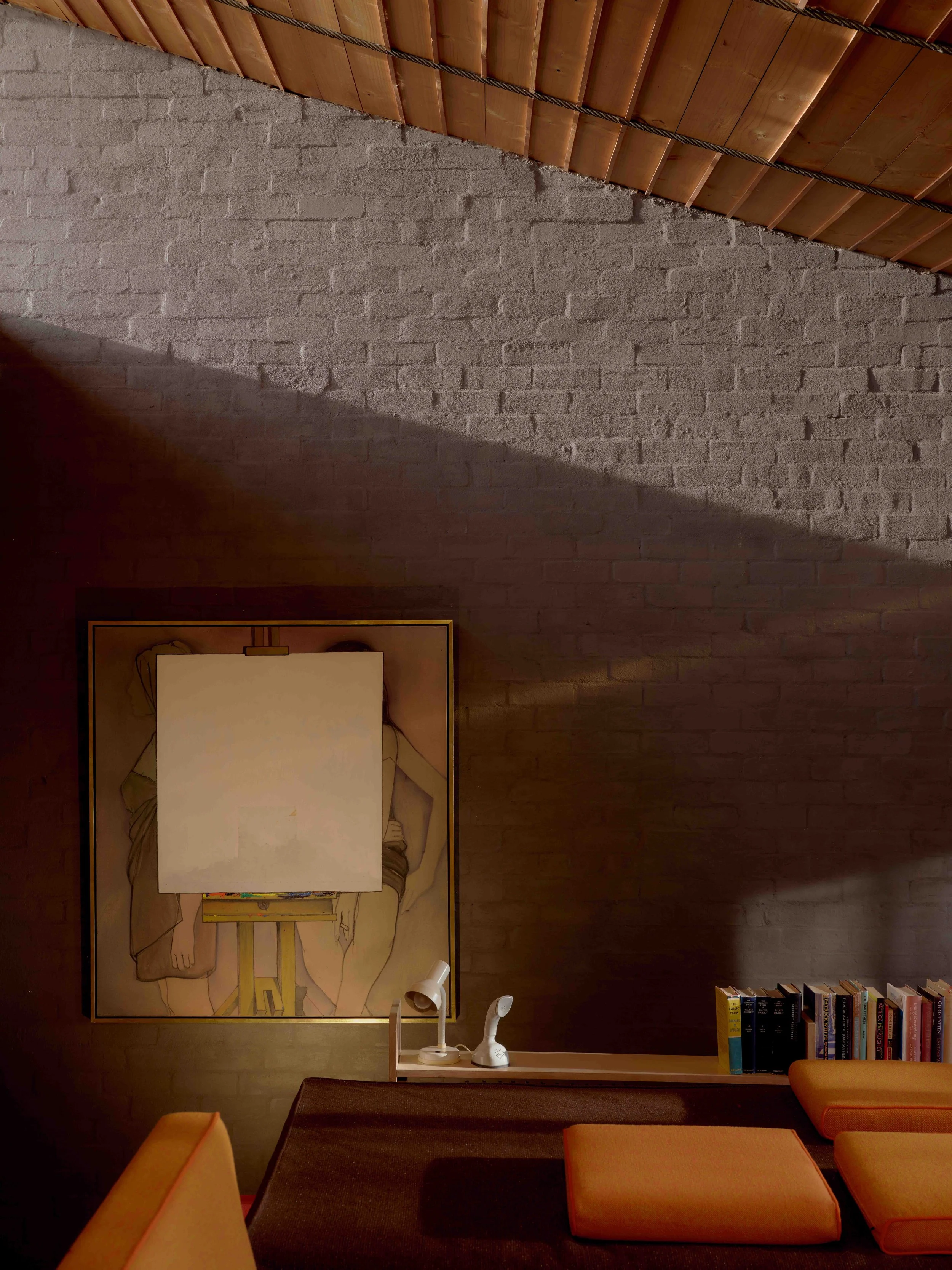 Interior view of a room with an exposed brick wall, a large framed artwork depicting a nude figure, a white adjustable desk lamp, a small white speaker, a wooden bookshelf with books, and a table with orange chairs and cushions.