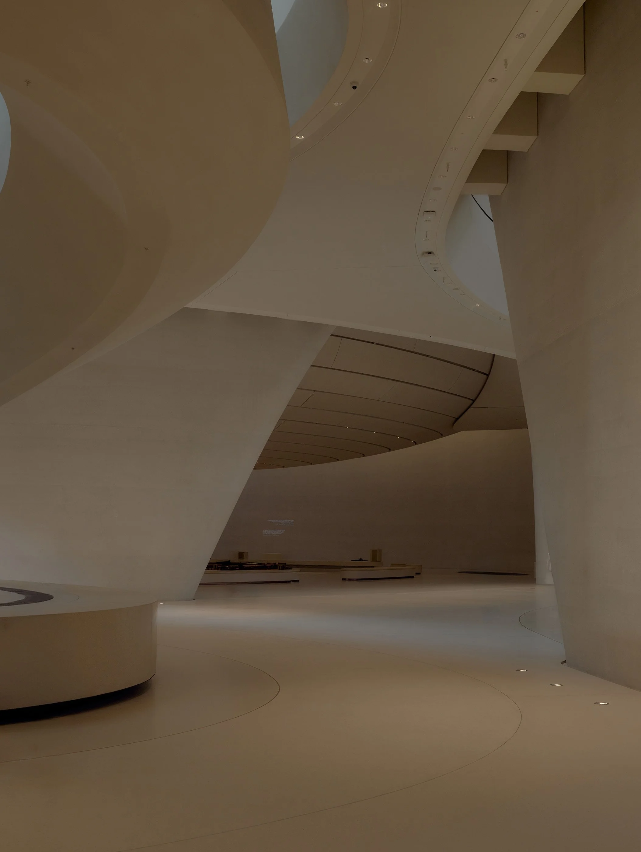 Abstract architectural interior with curved, flowing shapes in cream and beige tones, illuminated by recessed ceiling lights.
